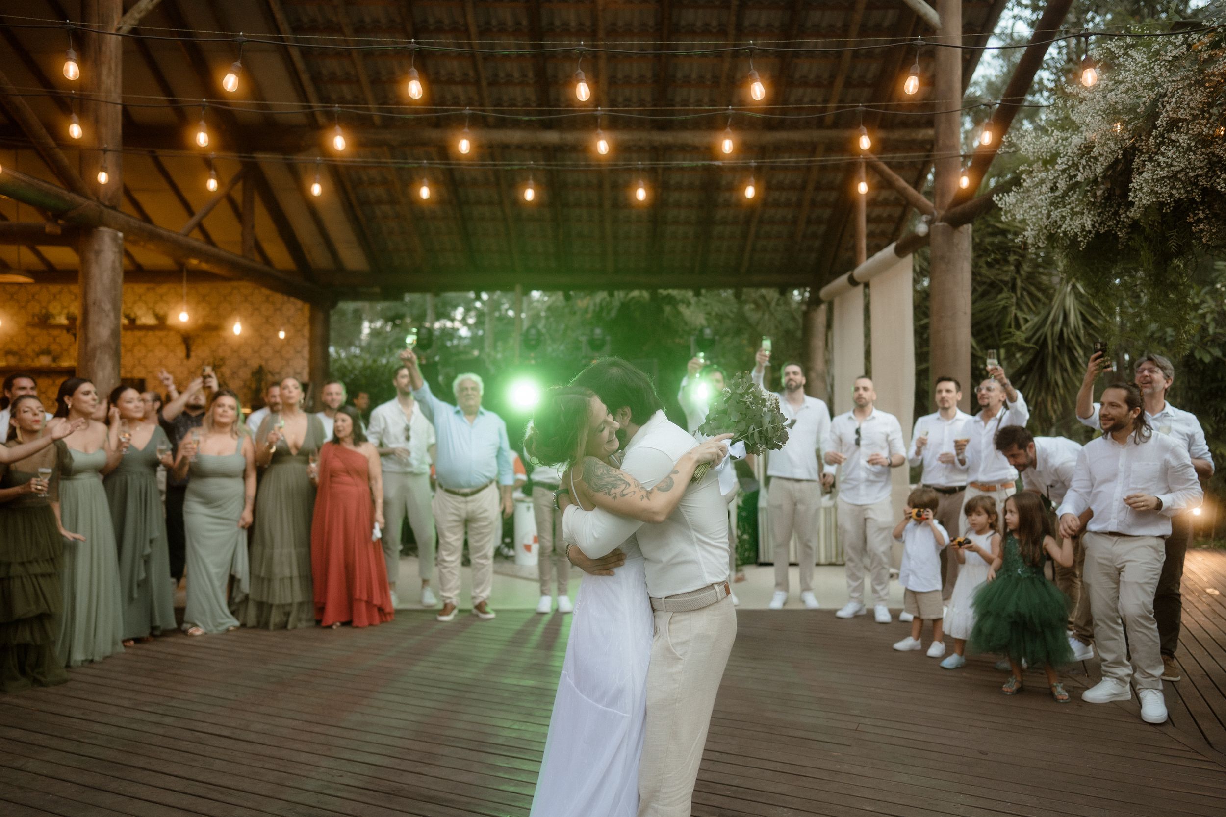 Newlyweds share their first dance surrounded by guests under string lights at an outdoor reception.