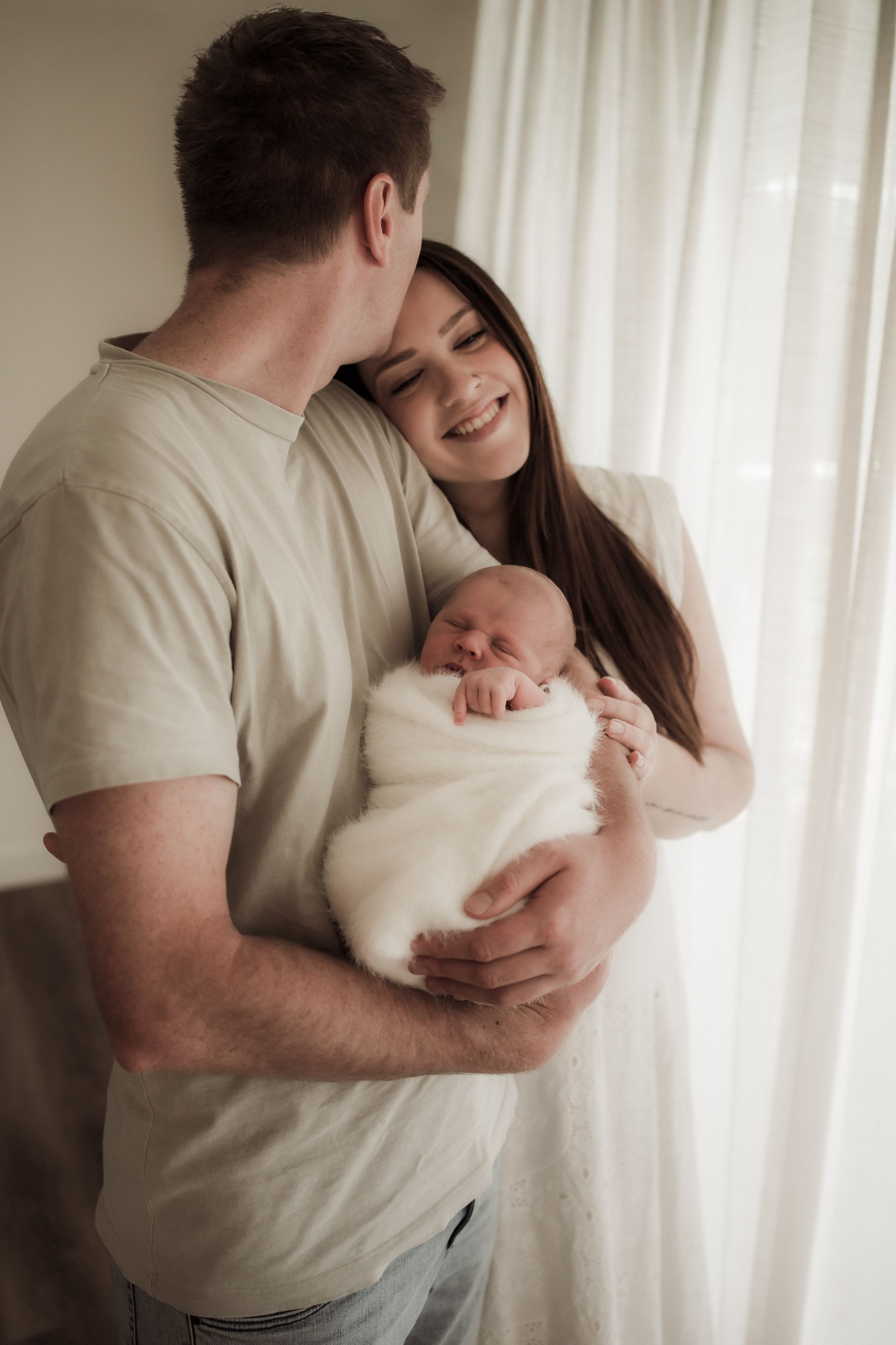 A couple stands by a window holding their newborn baby wrapped in white, sharing an intimate family moment.