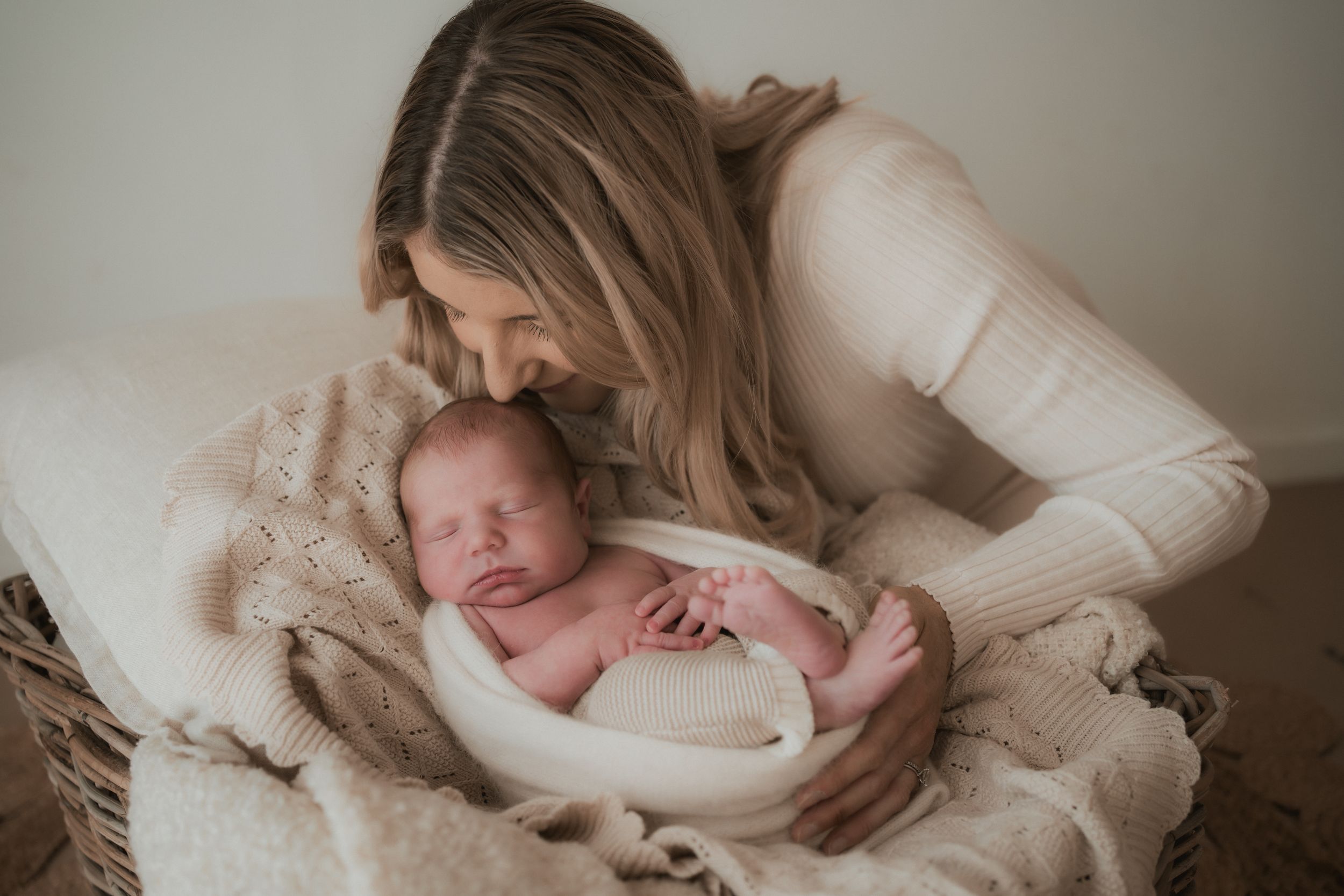 Newborn baby swaddled in soft cream blanket rests in woven basket during intimate portrait session.