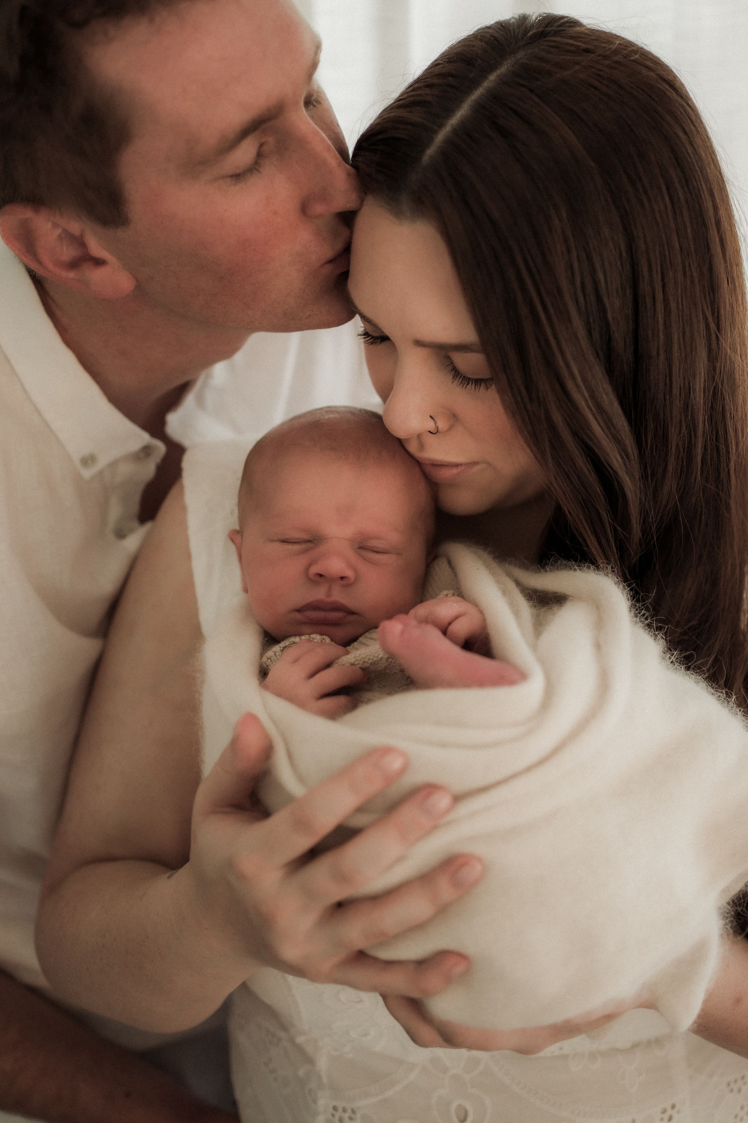 A series of tender moments showing parents cradling and kissing their sleeping newborn baby wrapped in white.