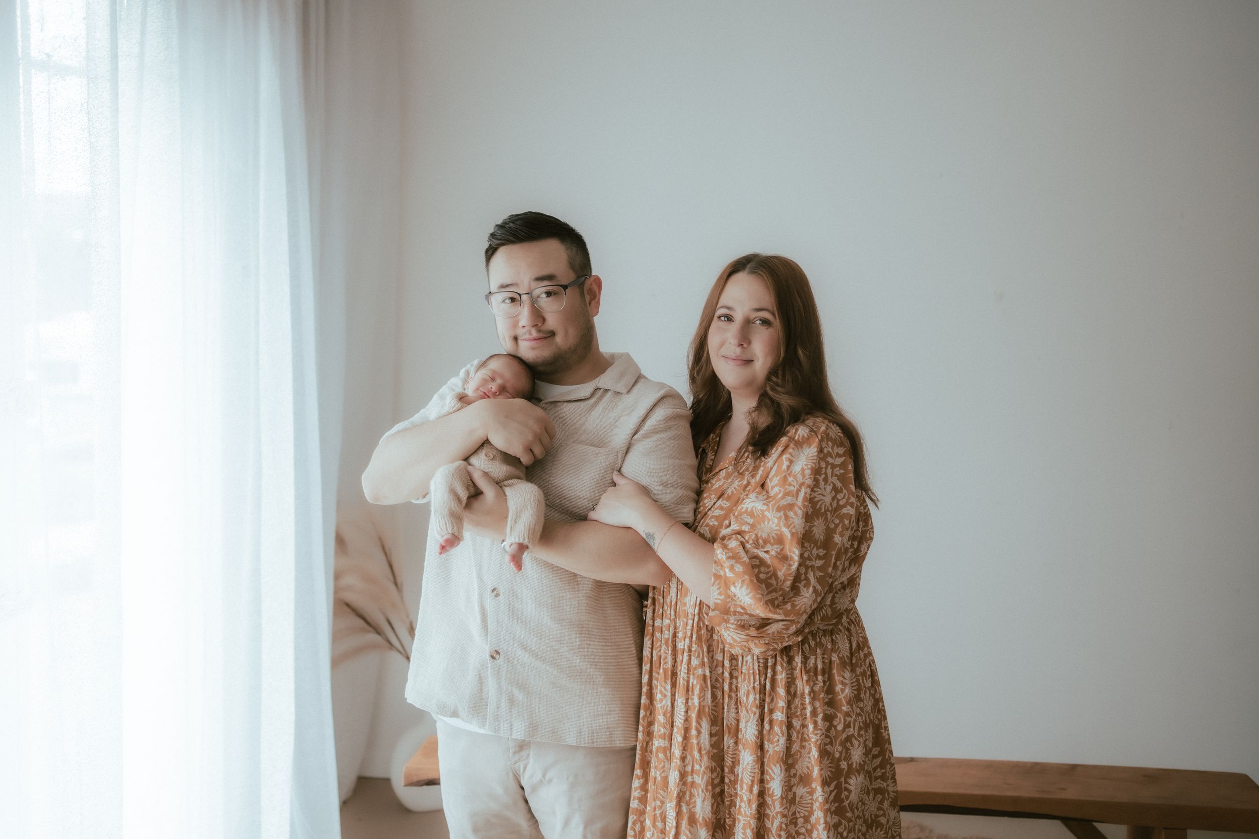 A couple in neutral tones stands near a bright window, with one wearing a floral dress and the other in beige attire.