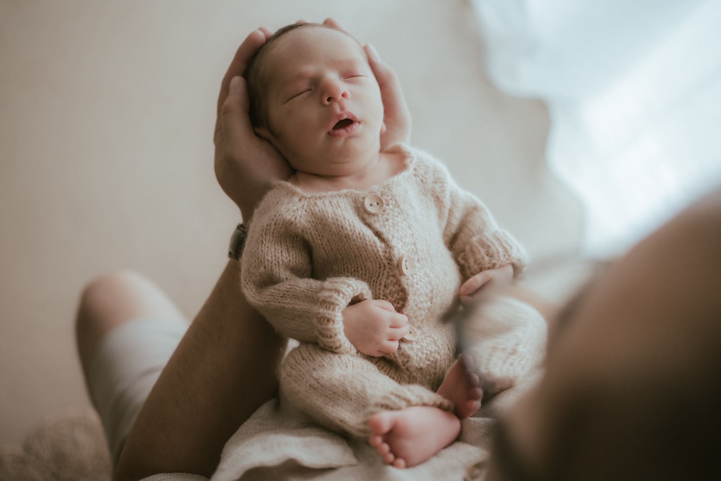 Sleeping newborn baby wearing cozy cream knit sweater rests peacefully while being held.