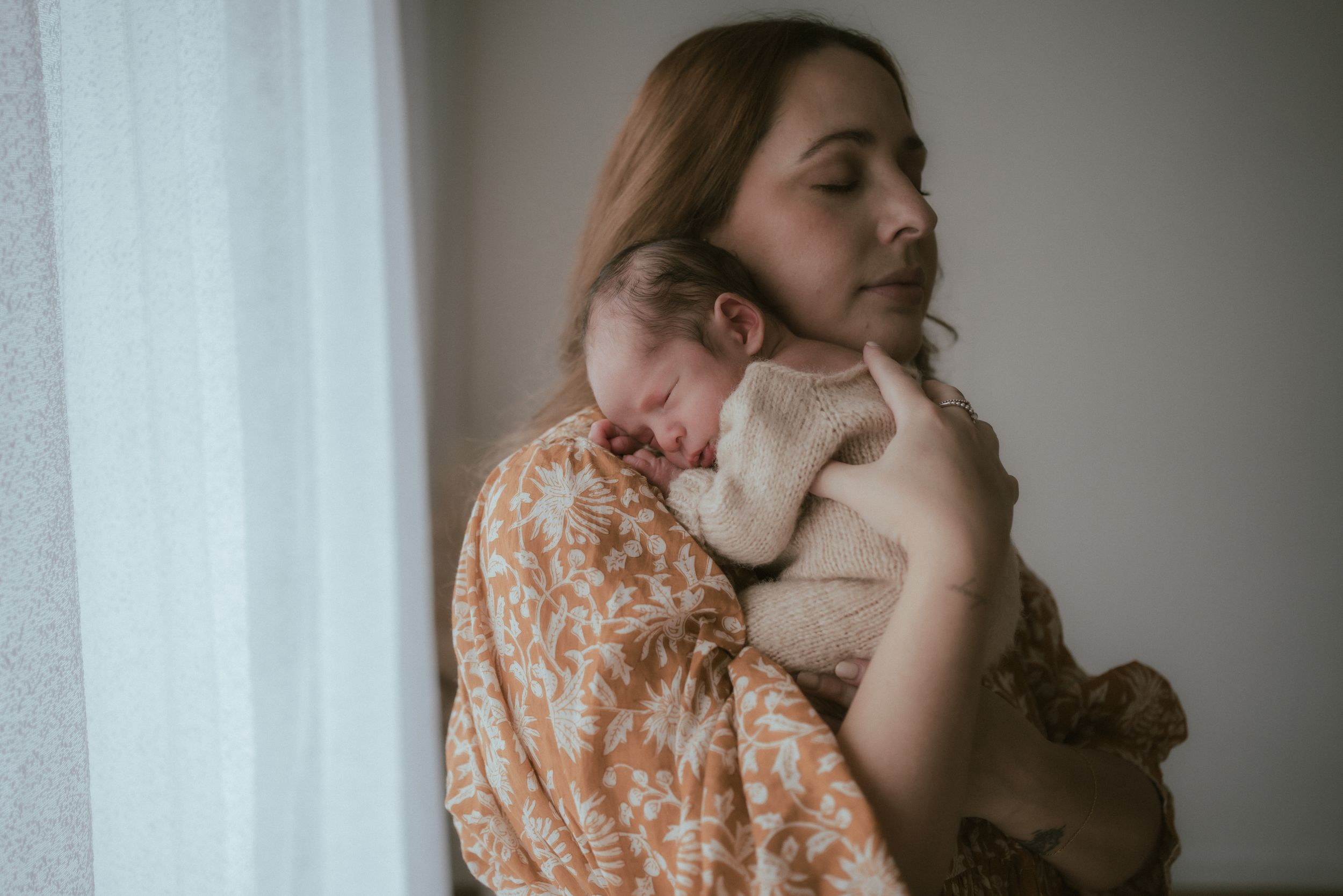 A person in a floral dress holds a sleeping newborn baby against their shoulder near a window.
