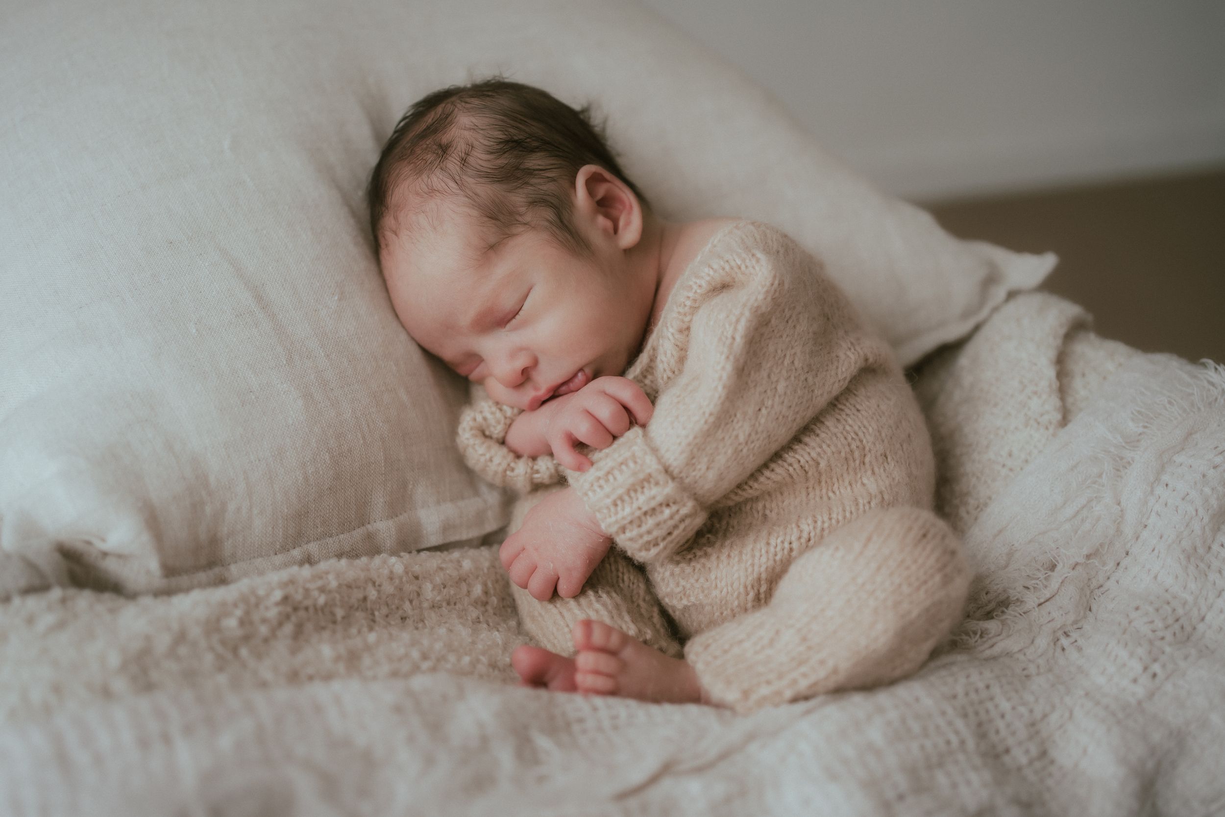 A sleeping newborn wearing a soft beige knit sweater curls up peacefully on white bedding.