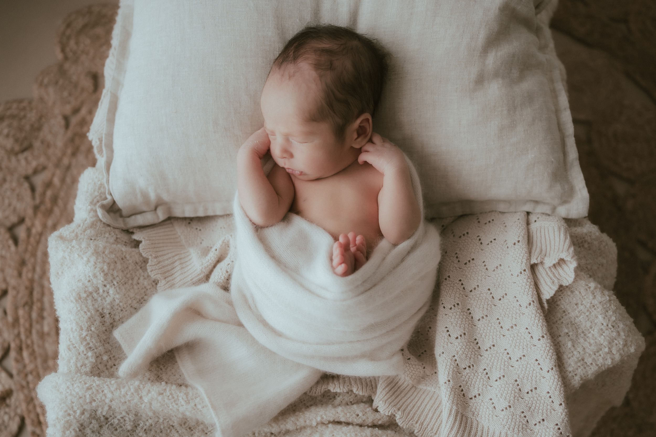 A sleeping newborn baby wrapped in white swaddle lies on soft textured bedding.