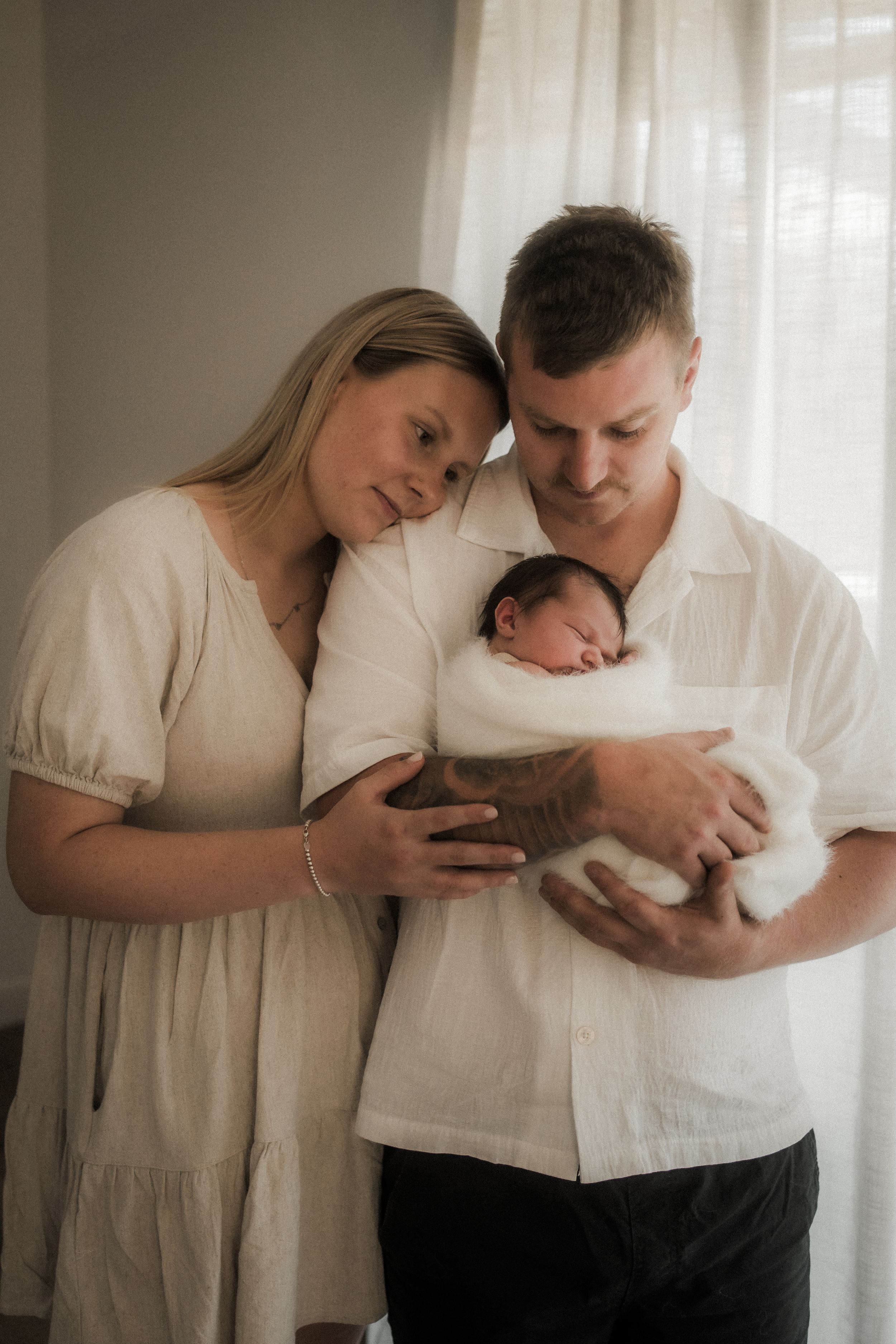 A couple in white clothing shares tender moments while holding their newborn baby in a bright indoor setting.