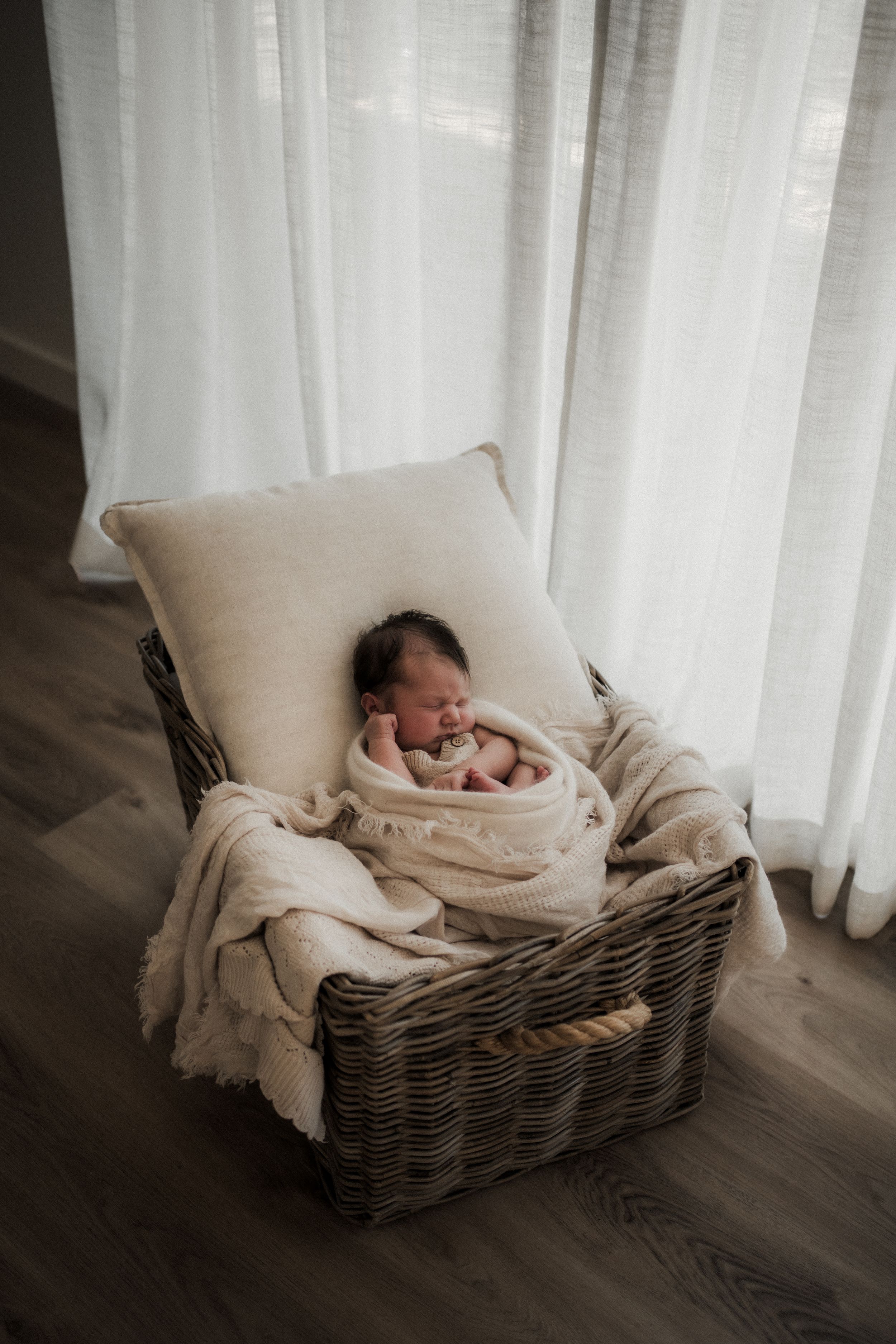 A newborn rests in a woven basket with cream-colored blankets near sheer white curtains.