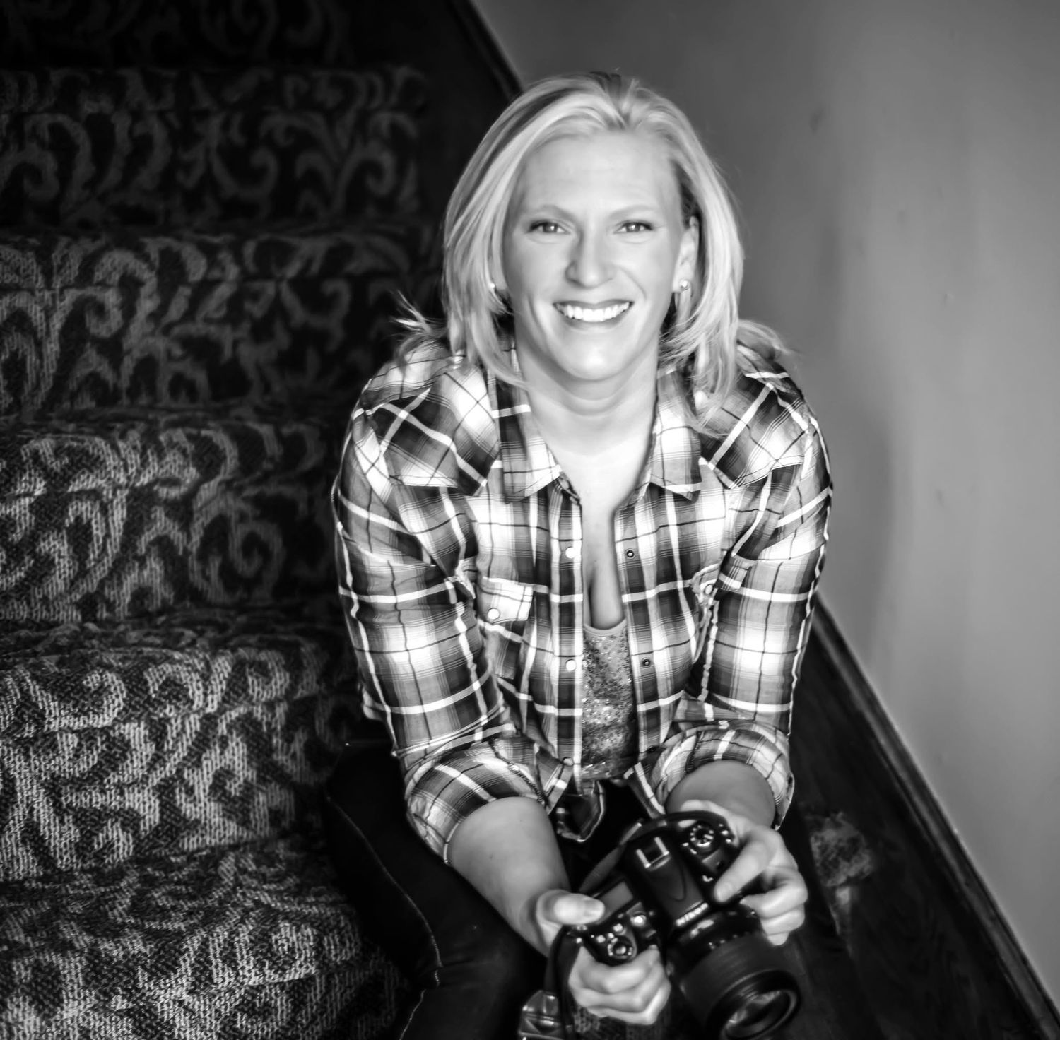 Black and white portrait of someone wearing a plaid shirt and holding a camera while sitting on patterned furniture.