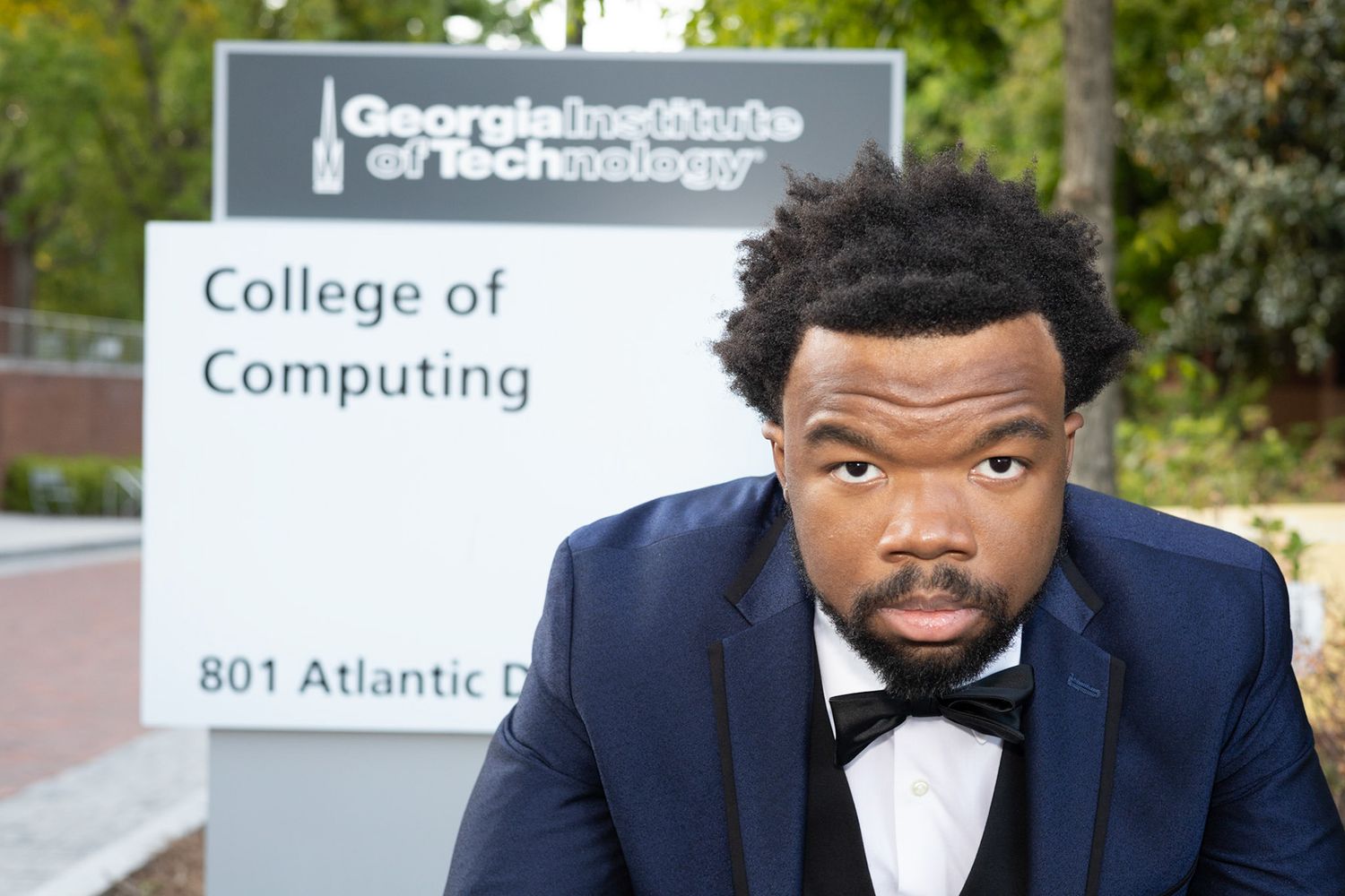 Graduate in navy suit and bowtie poses by College of Computing sign.