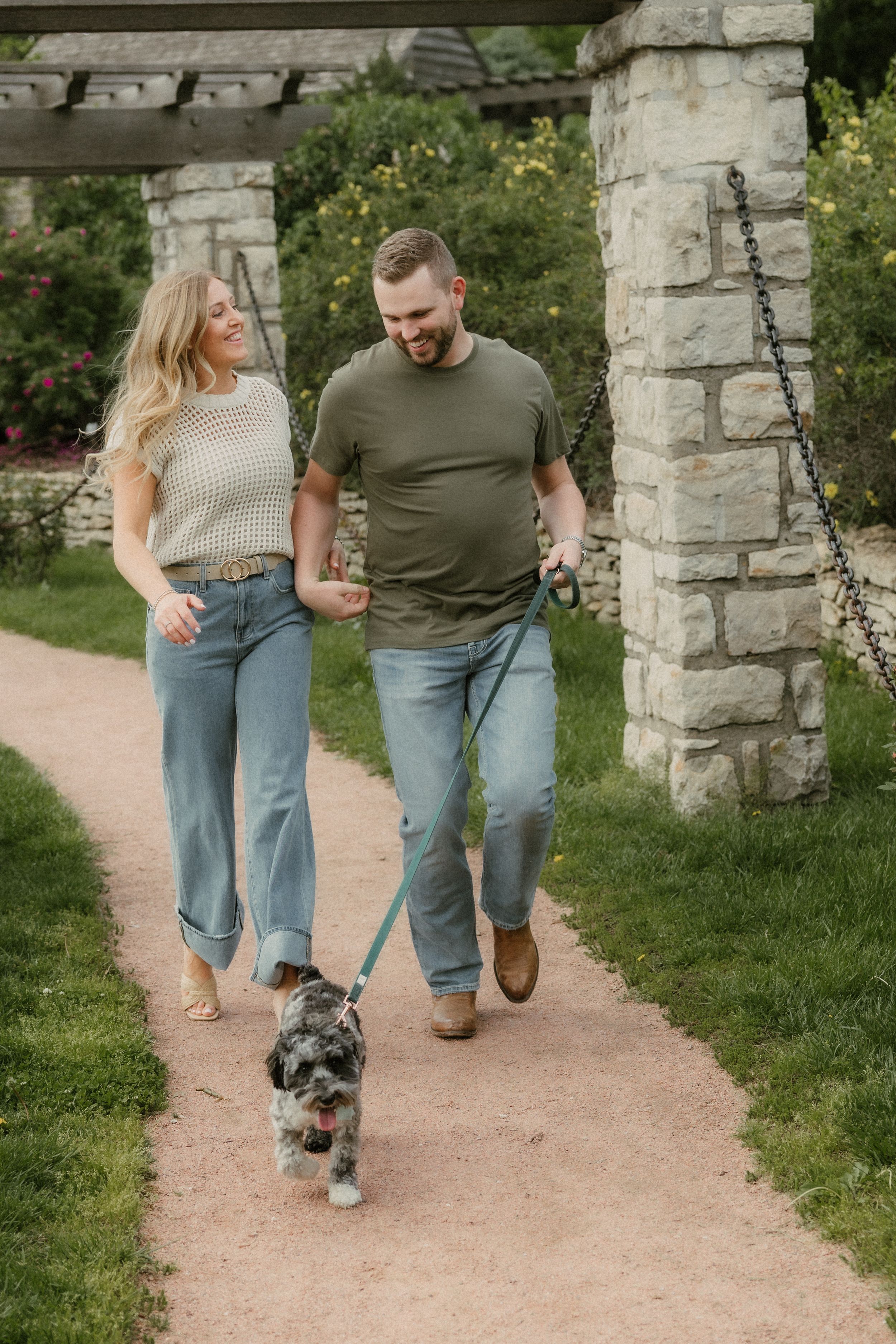Ben and Skylar laugh while walking hand in hand through Loose Park during their spring Kansas City engagement session with their dog trottin
