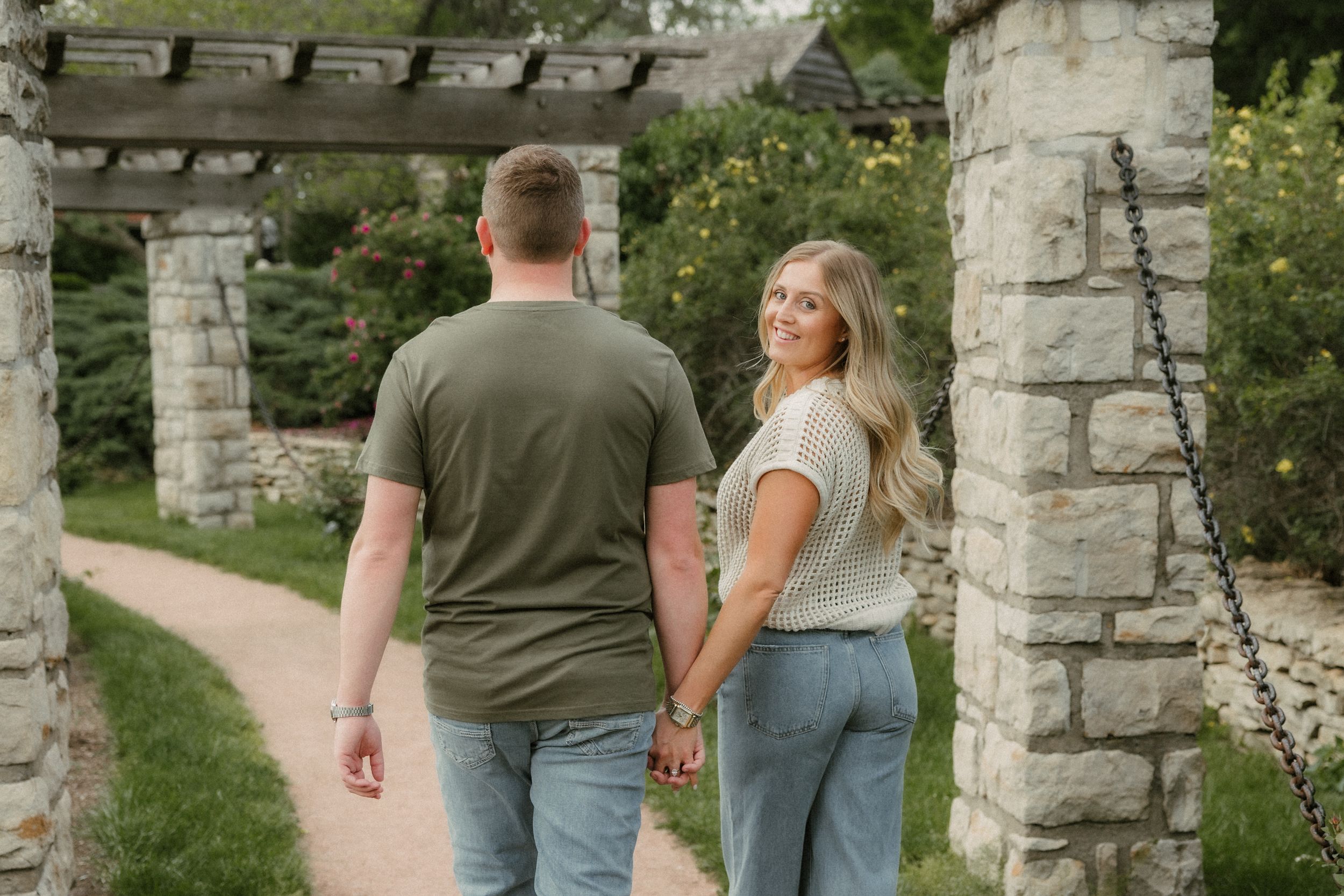 A couple walks together on a garden path between stone pillars and flowering plants on a sunny afternoon.