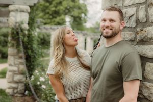 A couple stands close together by a stone wall outdoors, with one playfully teasing the other at Loose Park in Brookside Kansas.