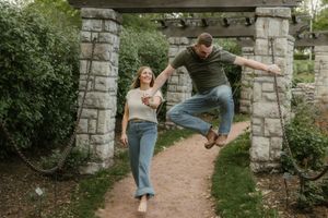 A playful couple poses at a stone archway entrance on a garden path while one person jumps mid-air at Kansas City’s Loose Park.