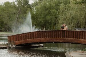 A couple embraces on a wooden footbridge over a lake with a fountain spraying in the background at Iconic Loose Park Bridge.