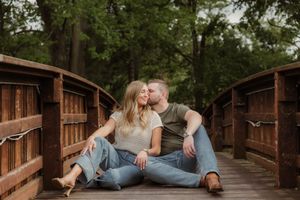 Two people in casual clothing sit close together on a wooden bridge surrounded by green trees in a romantic setting.