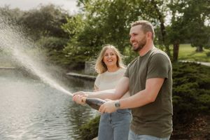 Couple spraying champagne and laughing together in a sunny garden setting near a pond at Loose Park.
