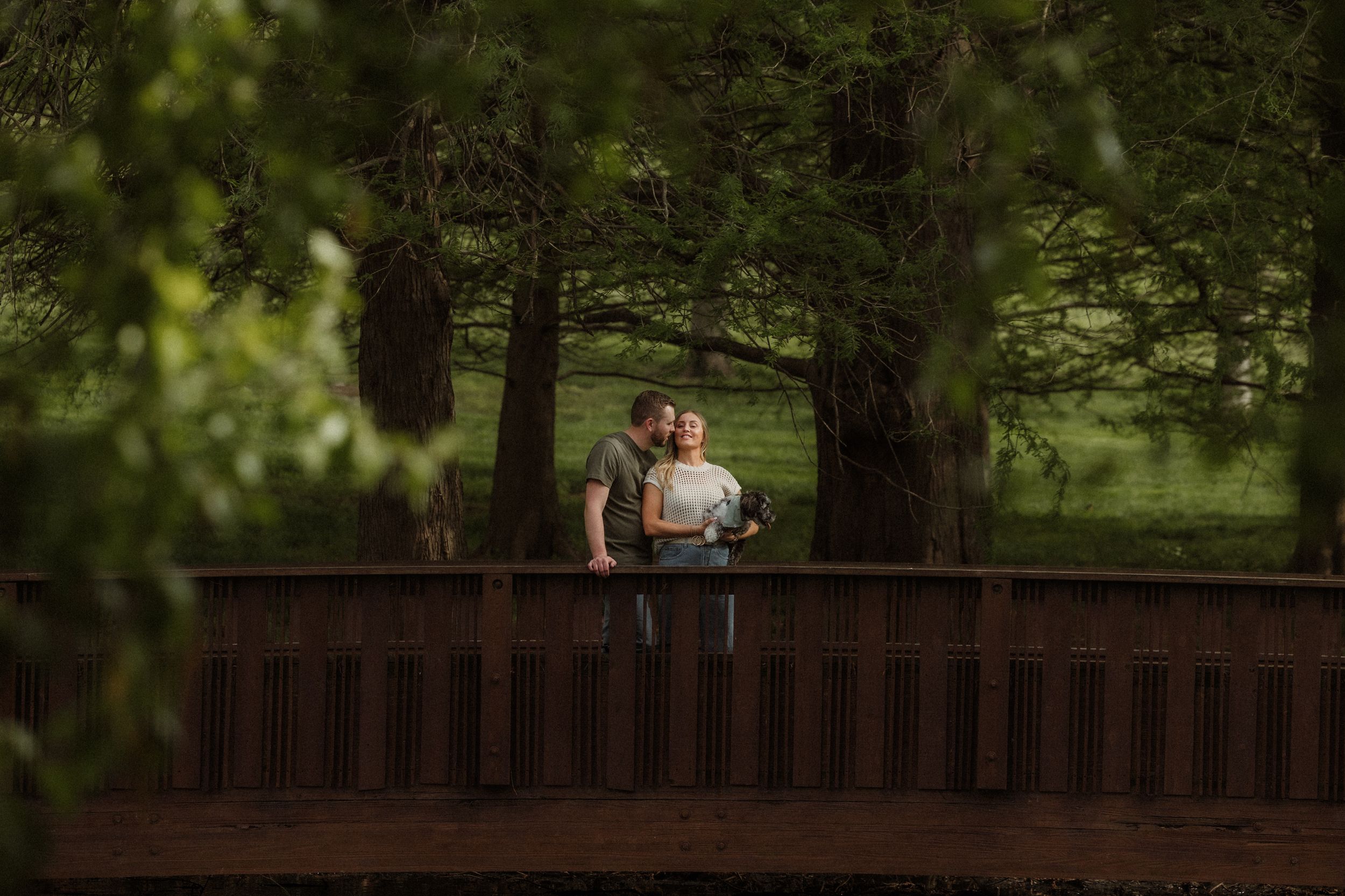 A couple stands together on a wooden bridge in a lush green  tree lined park setting during golden hour at Loose Park in Kansas City.