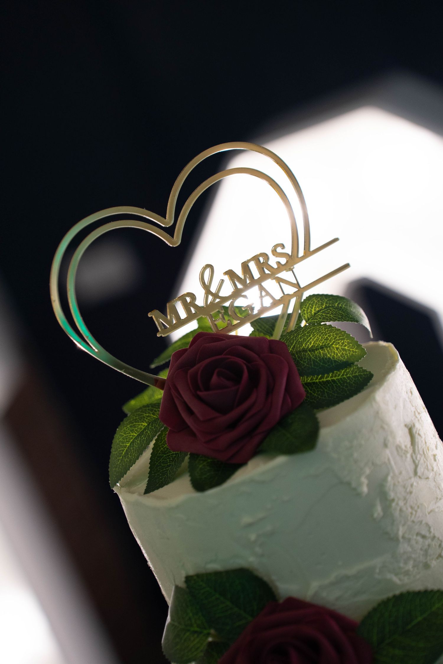 Wedding cake topper featuring a gold heart shape with Mr & Mrs text and burgundy roses against a dark background.