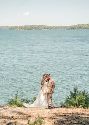 A couple stands together on a clifftop overlooking turquoise waters and distant wooded shoreline on their wedding day.