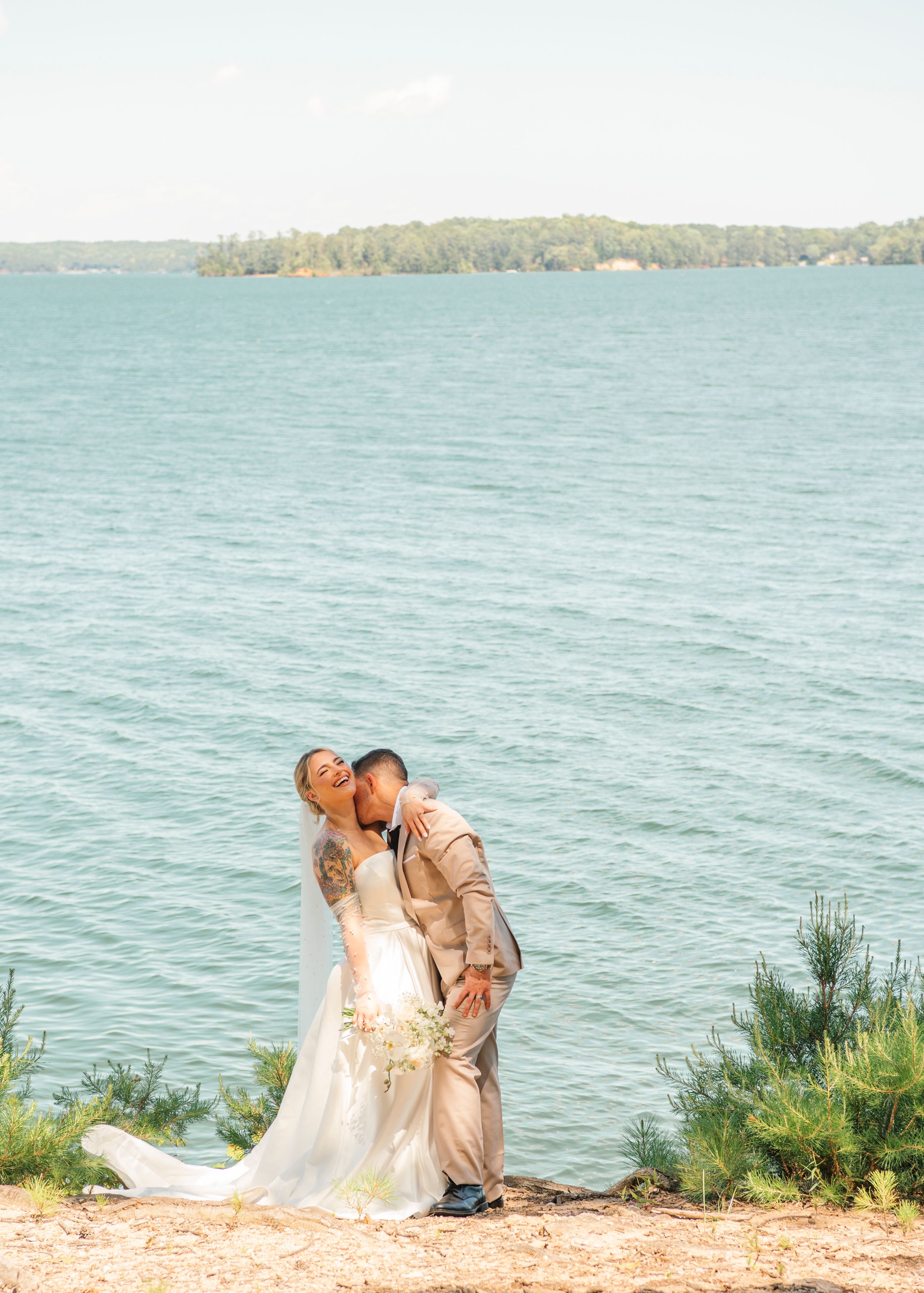 A couple shares an intimate moment on a lakeside beach with turquoise waters and wooded shoreline in the background.