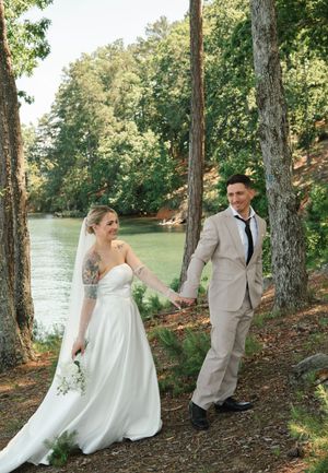A couple in wedding attire holds hands while standing on a wooded path beside a tranquil lake on their special day.