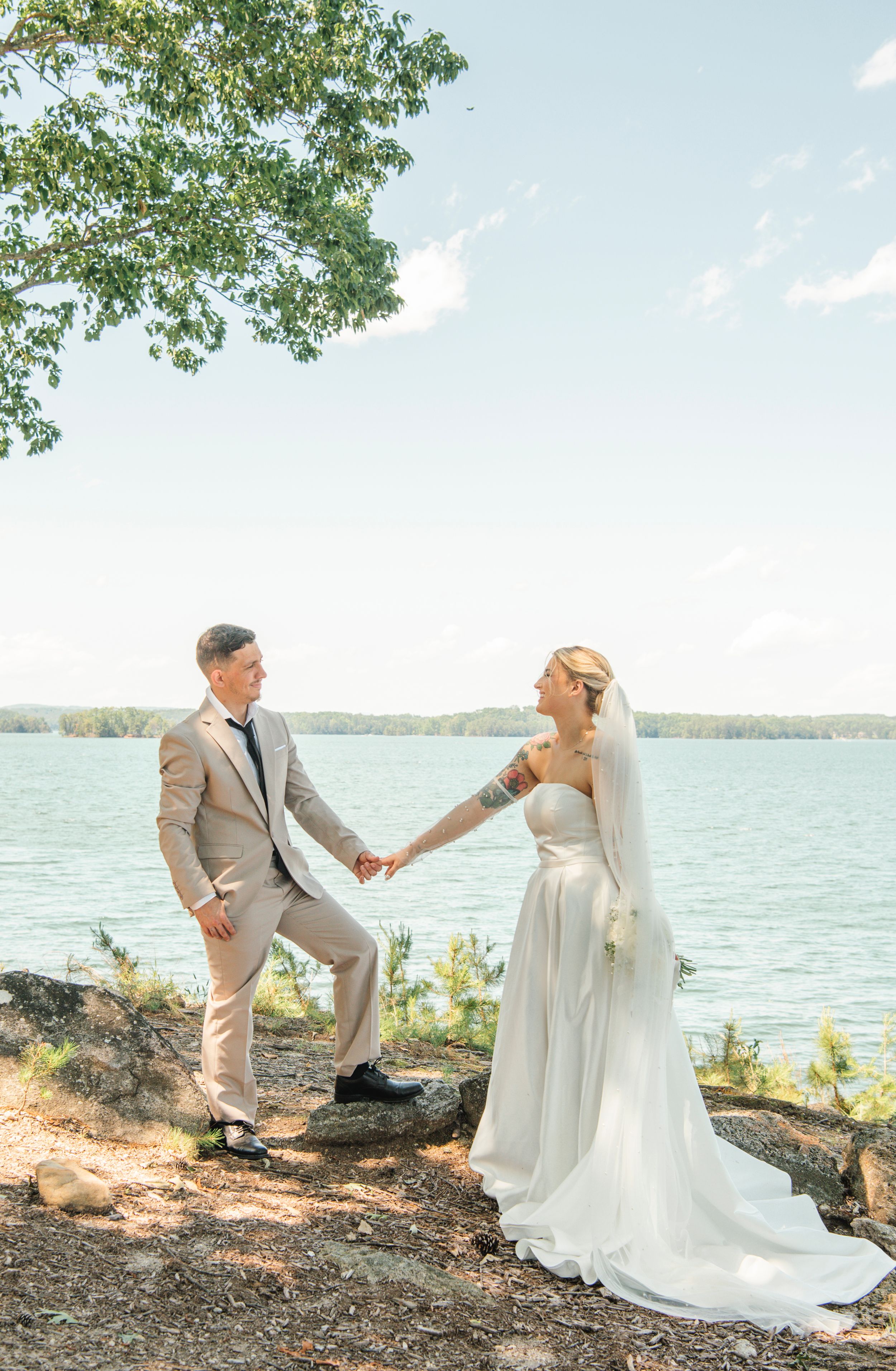 A couple poses in elegant wedding attire on rocky terrain overlooking a serene lake on their special day.