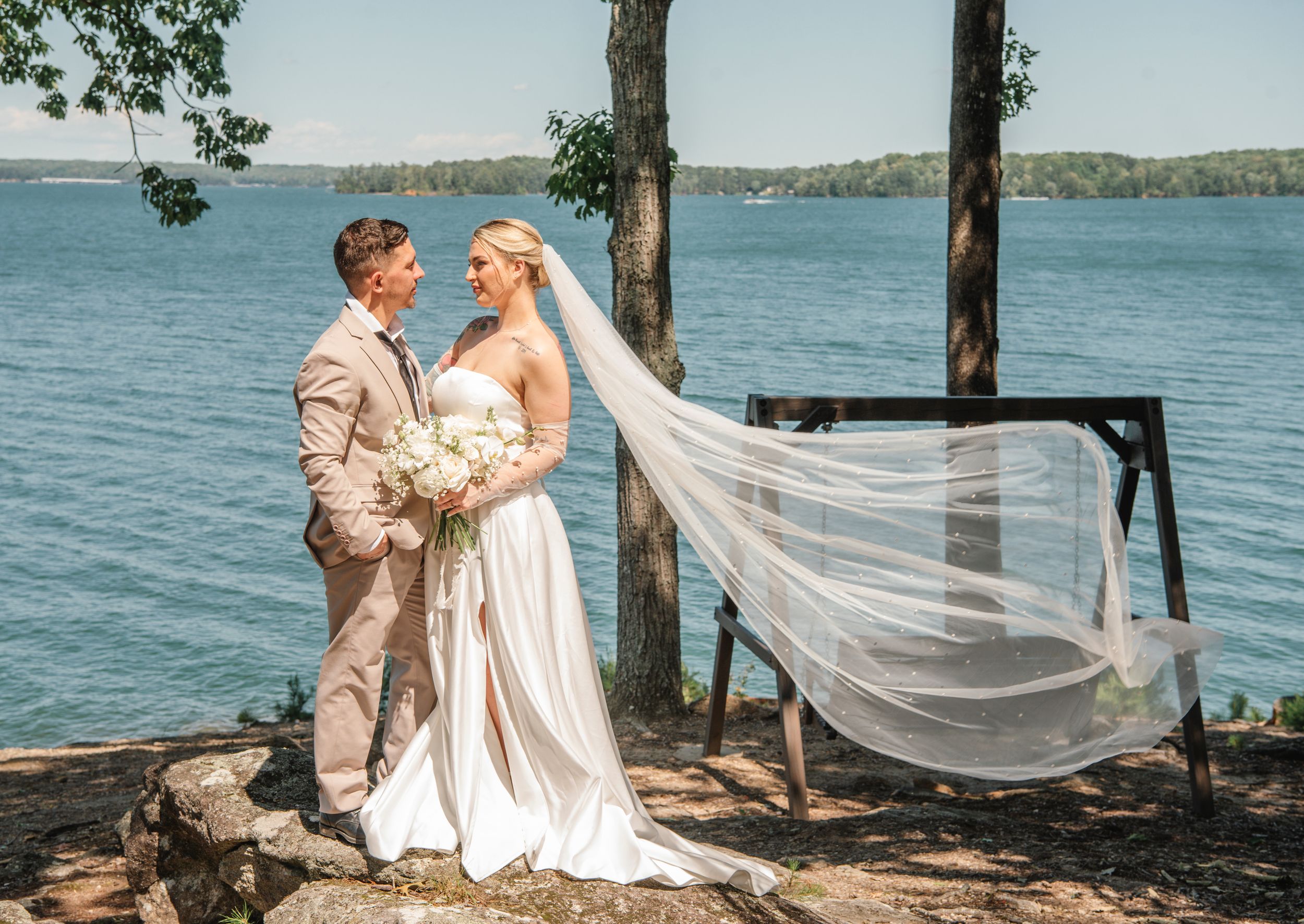 A romantic wedding couple poses on a lakeside shore with a flowing veil and tan suit against the water backdrop.