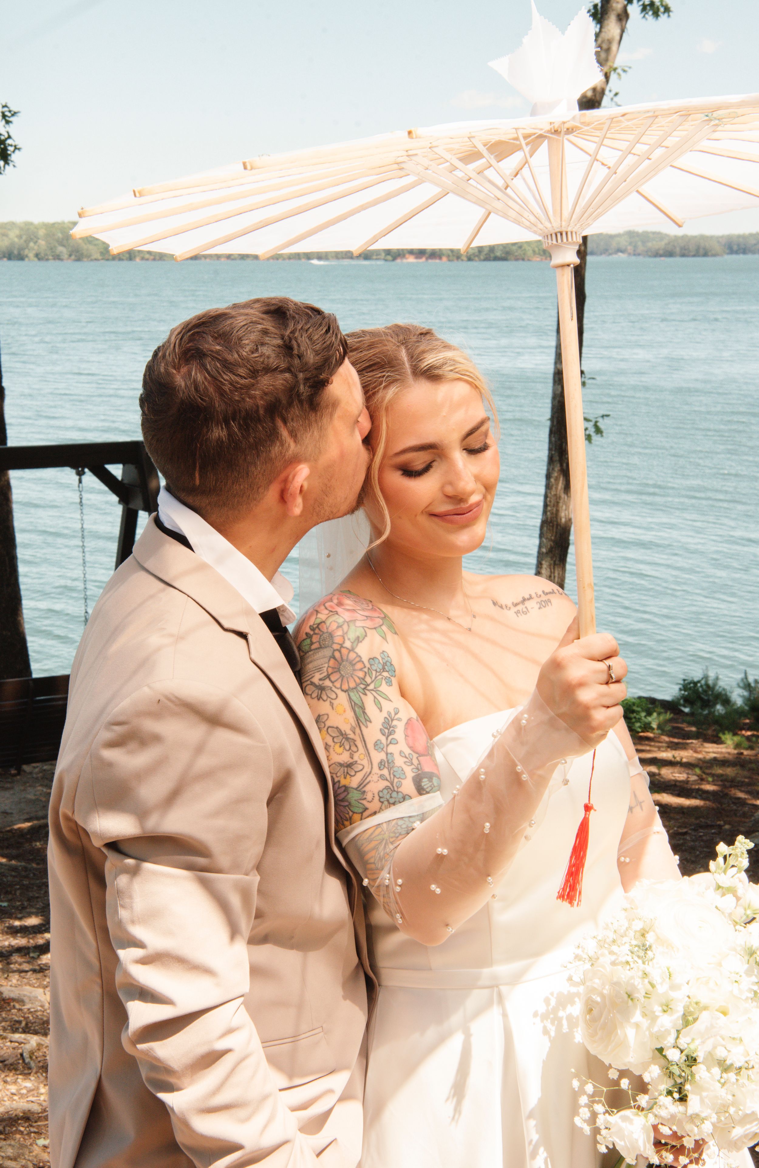 A couple shares an intimate moment under a white parasol by the waterfront on a sunny day.