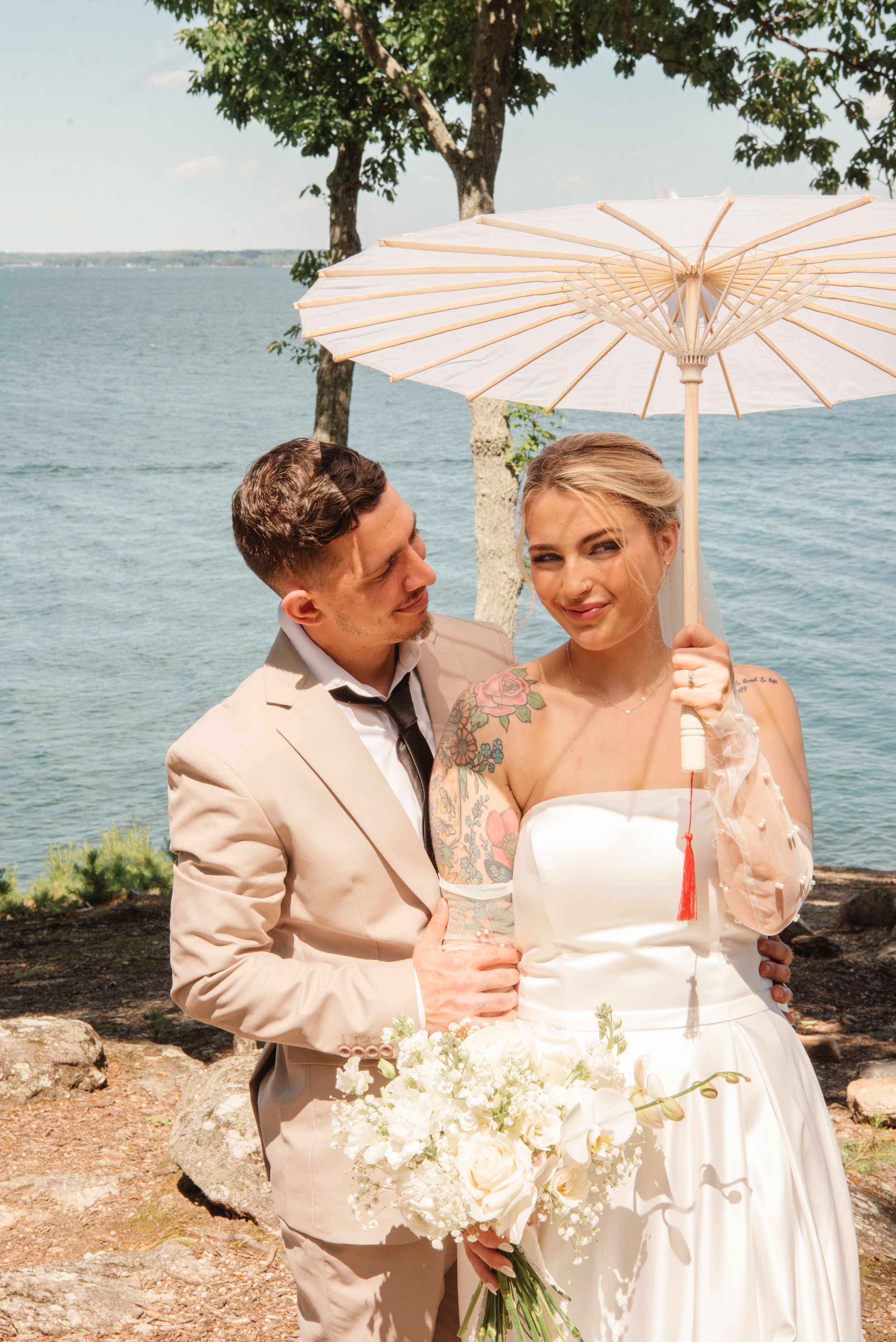 Wedding couple poses near waterfront with striped parasol and bouquet of white flowers on a sunny day.