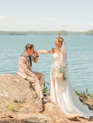 A couple shares an intimate moment on a rocky lakeside cliff with the bride in white and groom in beige suit.