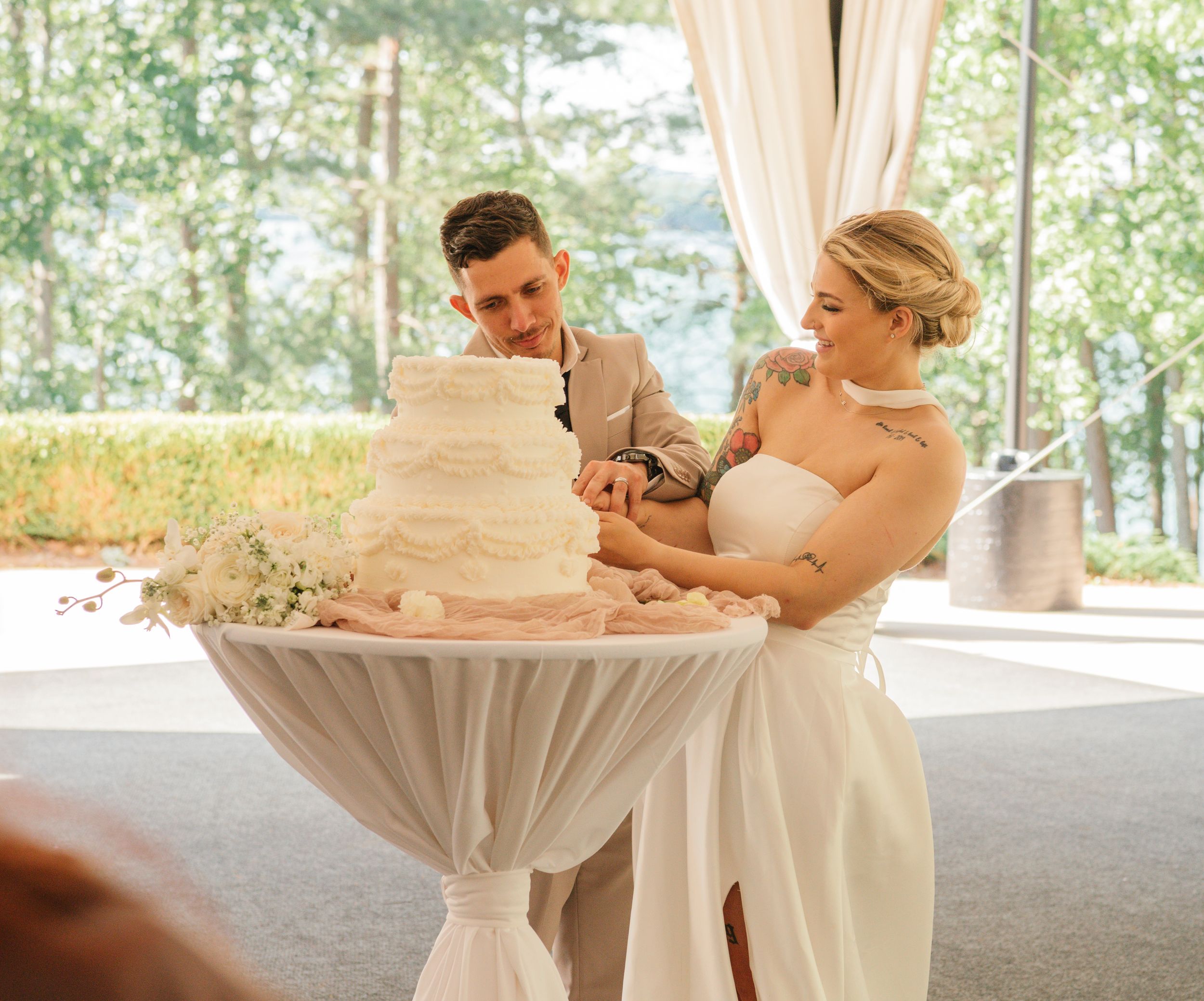 A couple cutting their elegant three-tiered white wedding cake together at an outdoor reception venue.