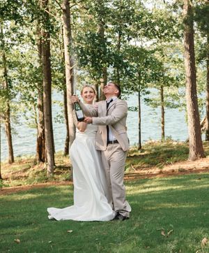 A couple embraces joyfully in their wedding attire on a grassy lawn with pine trees and a lake in the background.