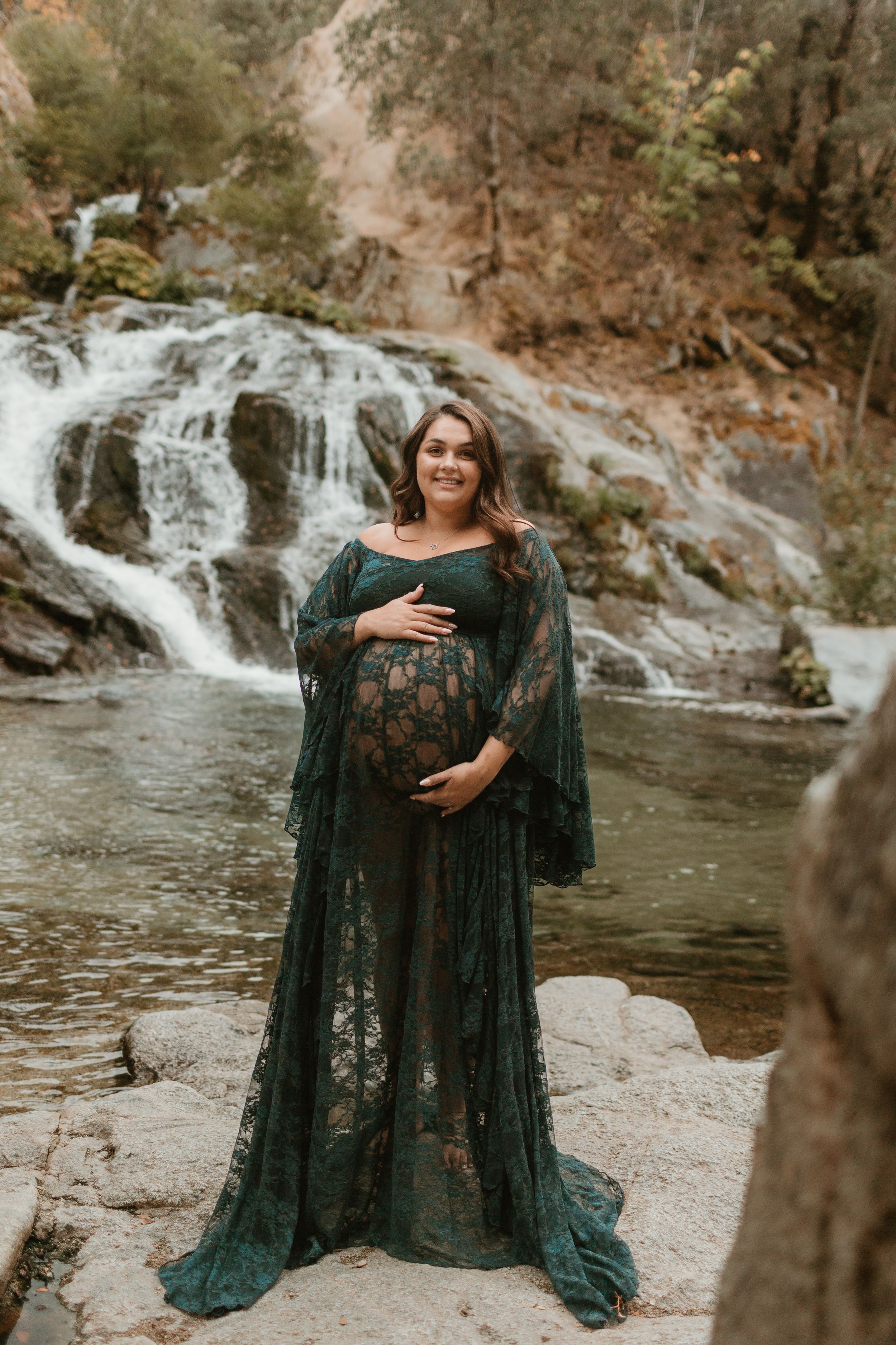 A flowing black lace dress is showcased against a scenic waterfall backdrop in a dramatic maternity photoshoot.