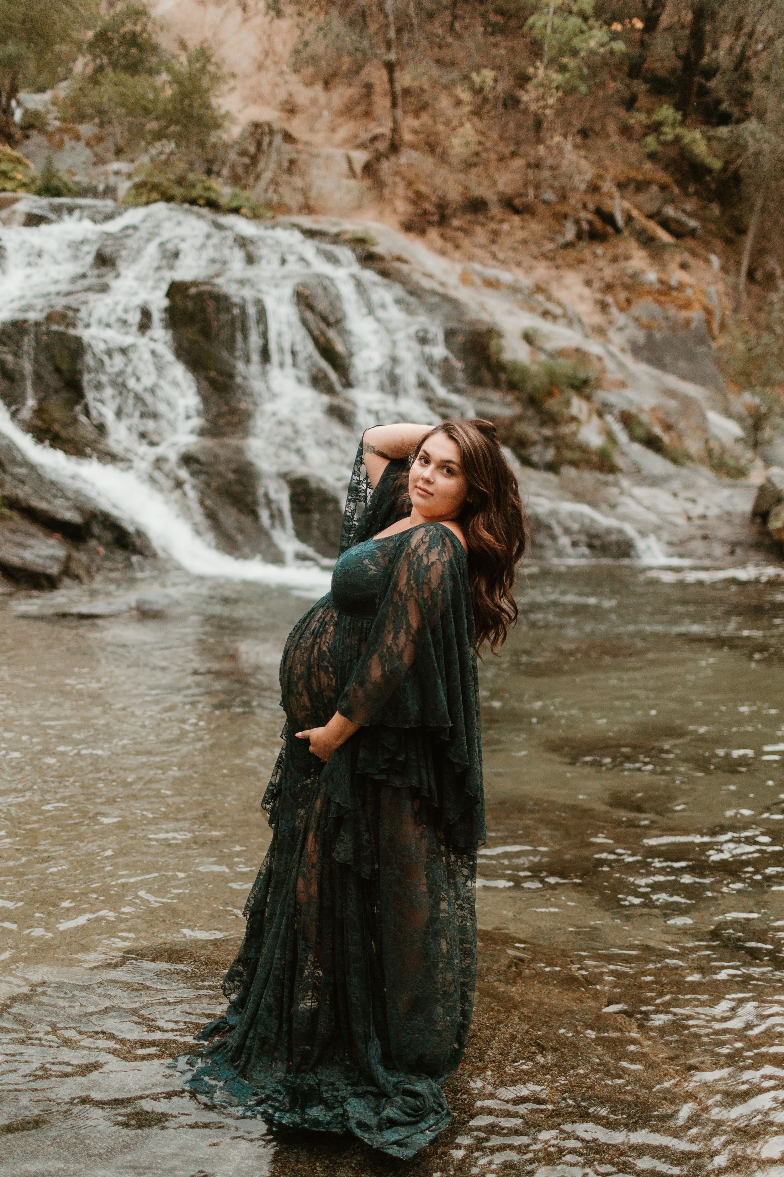 Artistic maternity photos featuring a black lace gown by a cascading waterfall in nature.