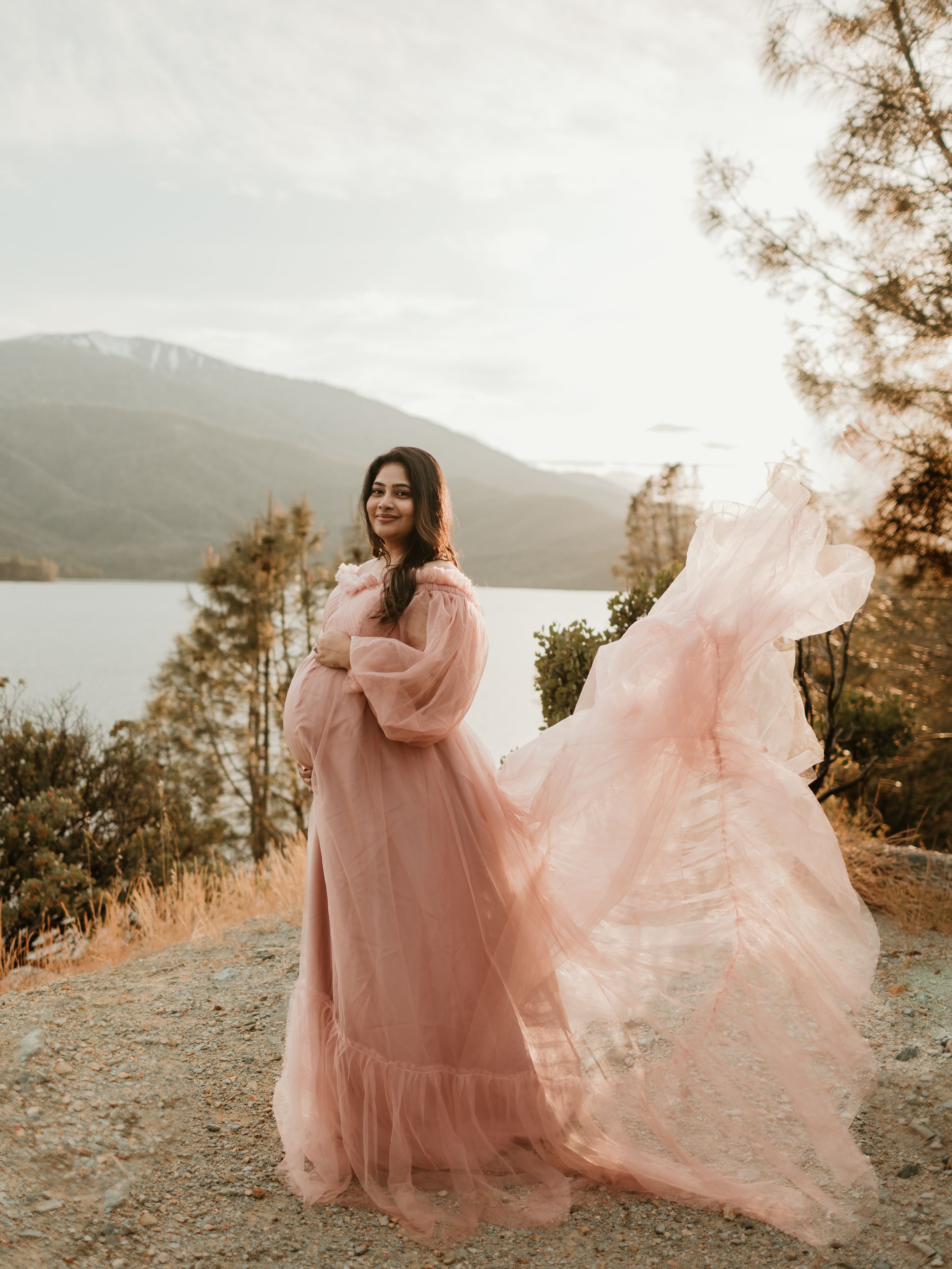 A flowing pink tulle dress catches the wind against a scenic mountain lake landscape at sunset.