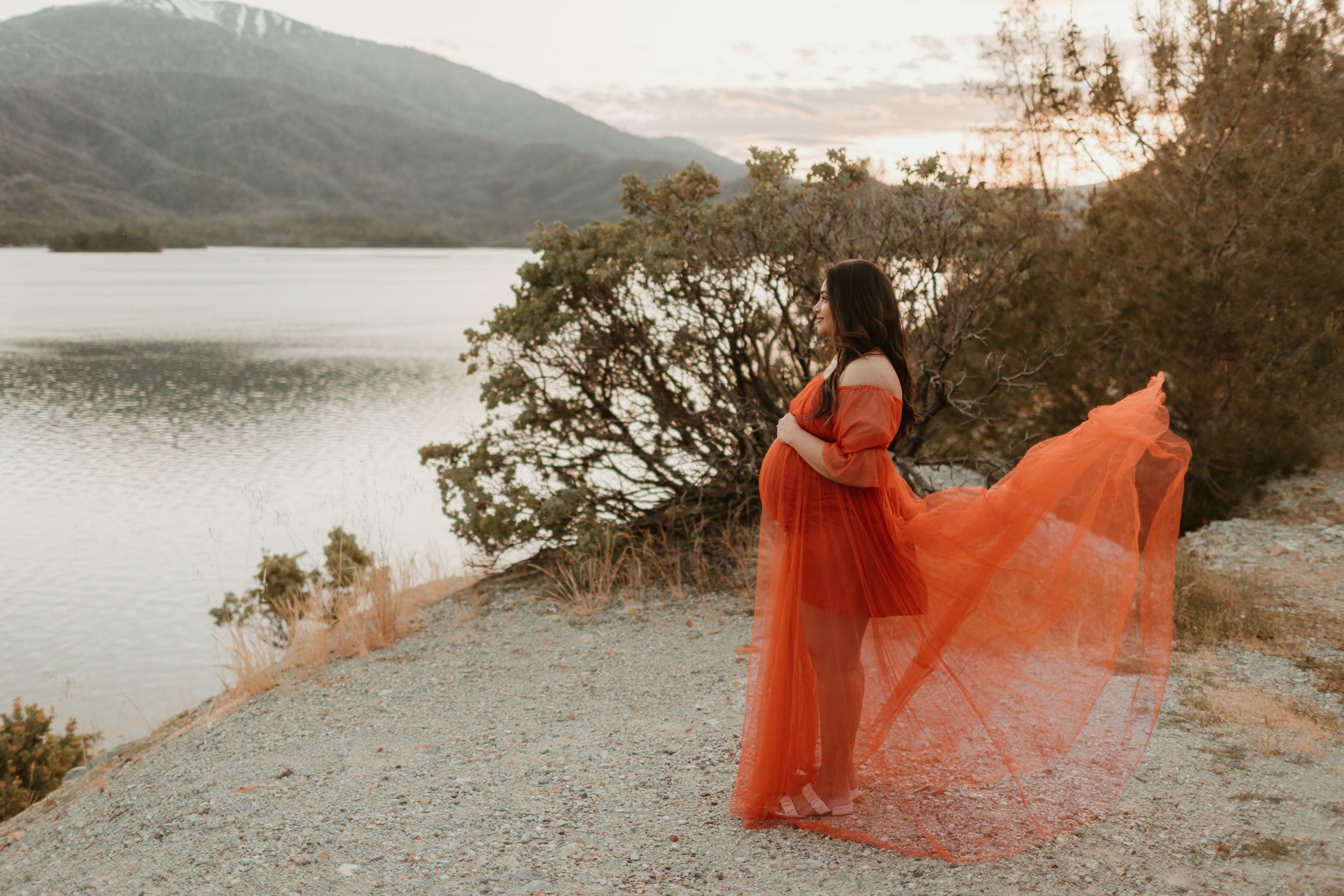 An orange-red dress billows dramatically against a rocky mountain landscape overlooking a serene lake.
