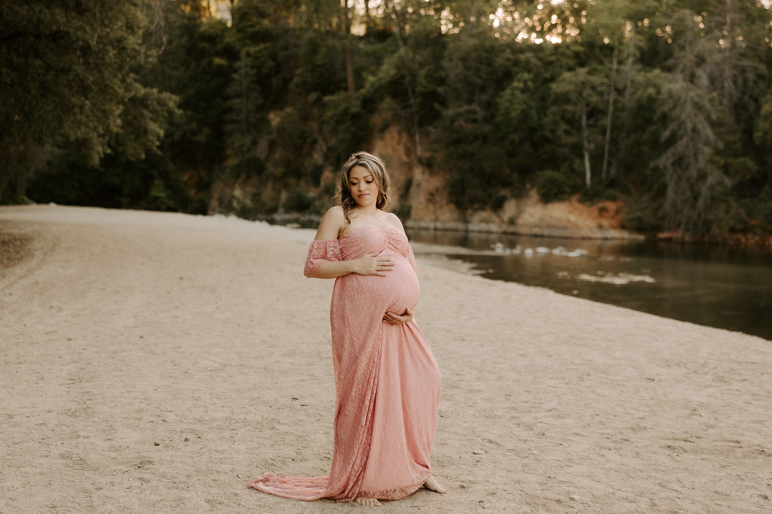 Maternity photoshoot on a sandy beach featuring a pink flowing dress at sunset.