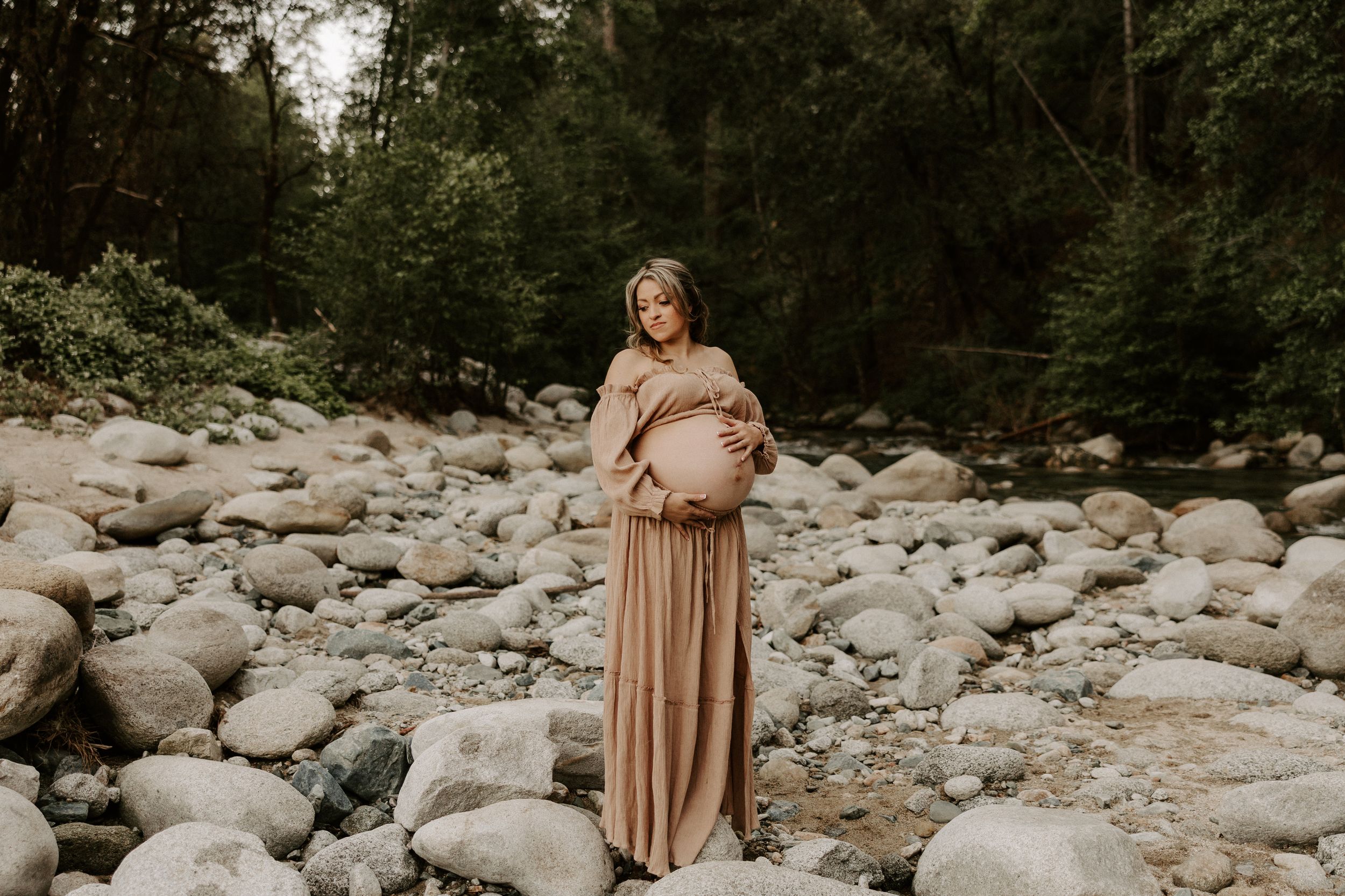 Maternity session among rocky riverbed terrain with flowing mauve dress.