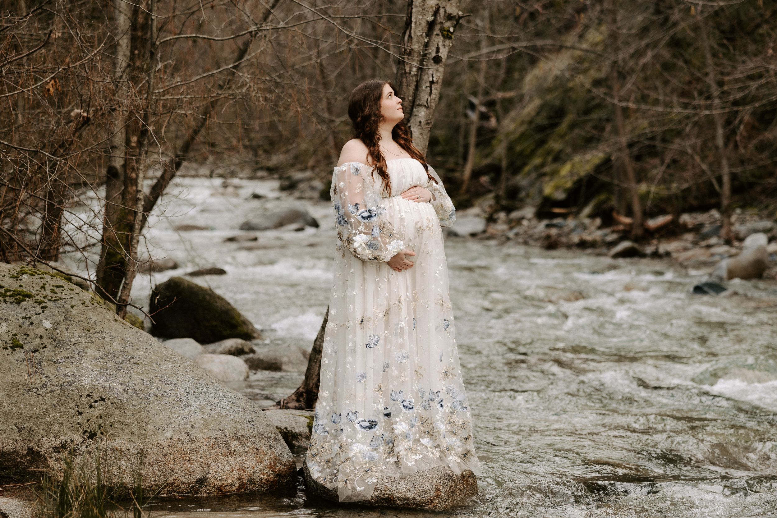 Ethereal white lace dress flows gracefully across rocky creek bed in moody forest setting.