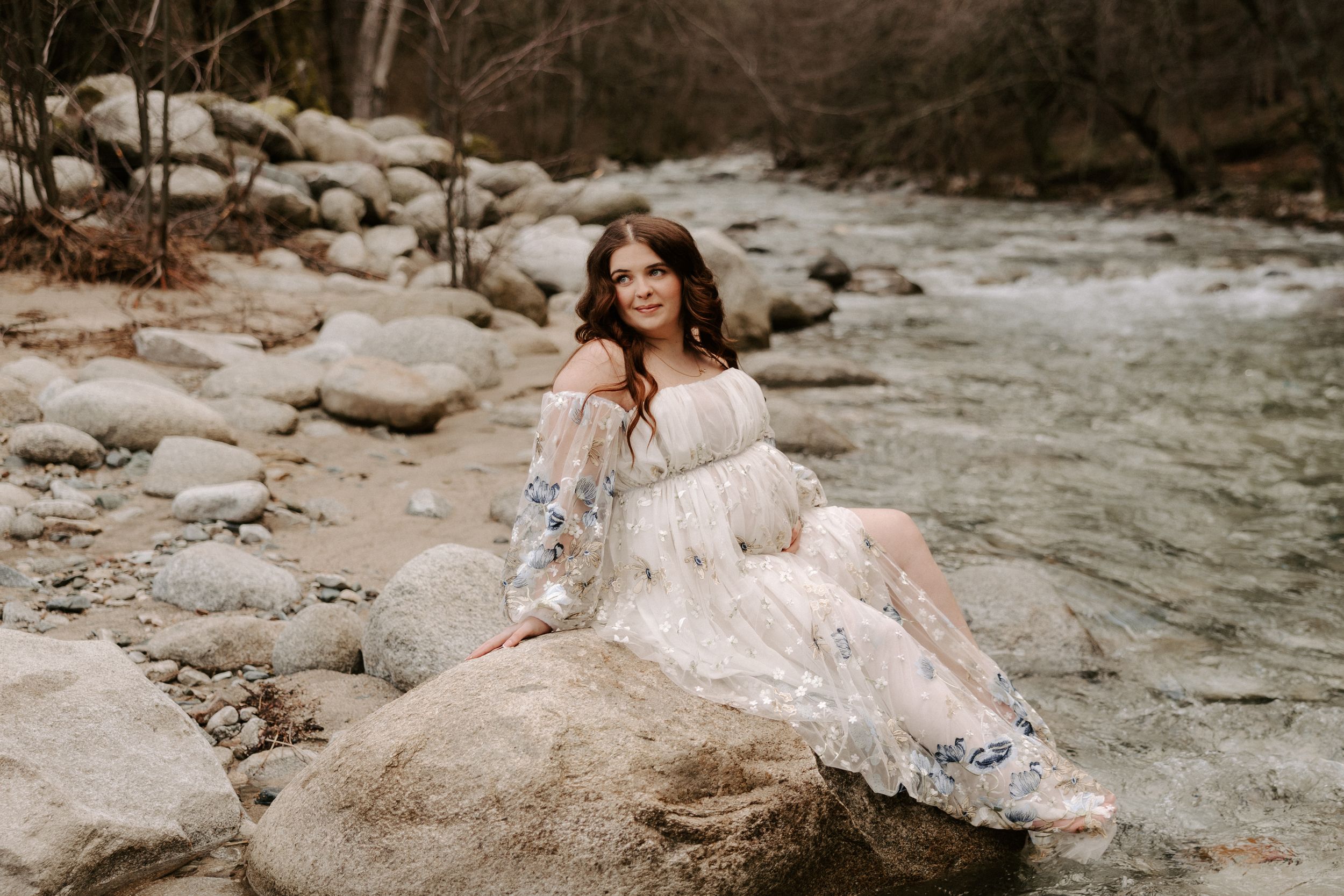 Elegant lace gown drapes over natural stone formations by a serene creek.