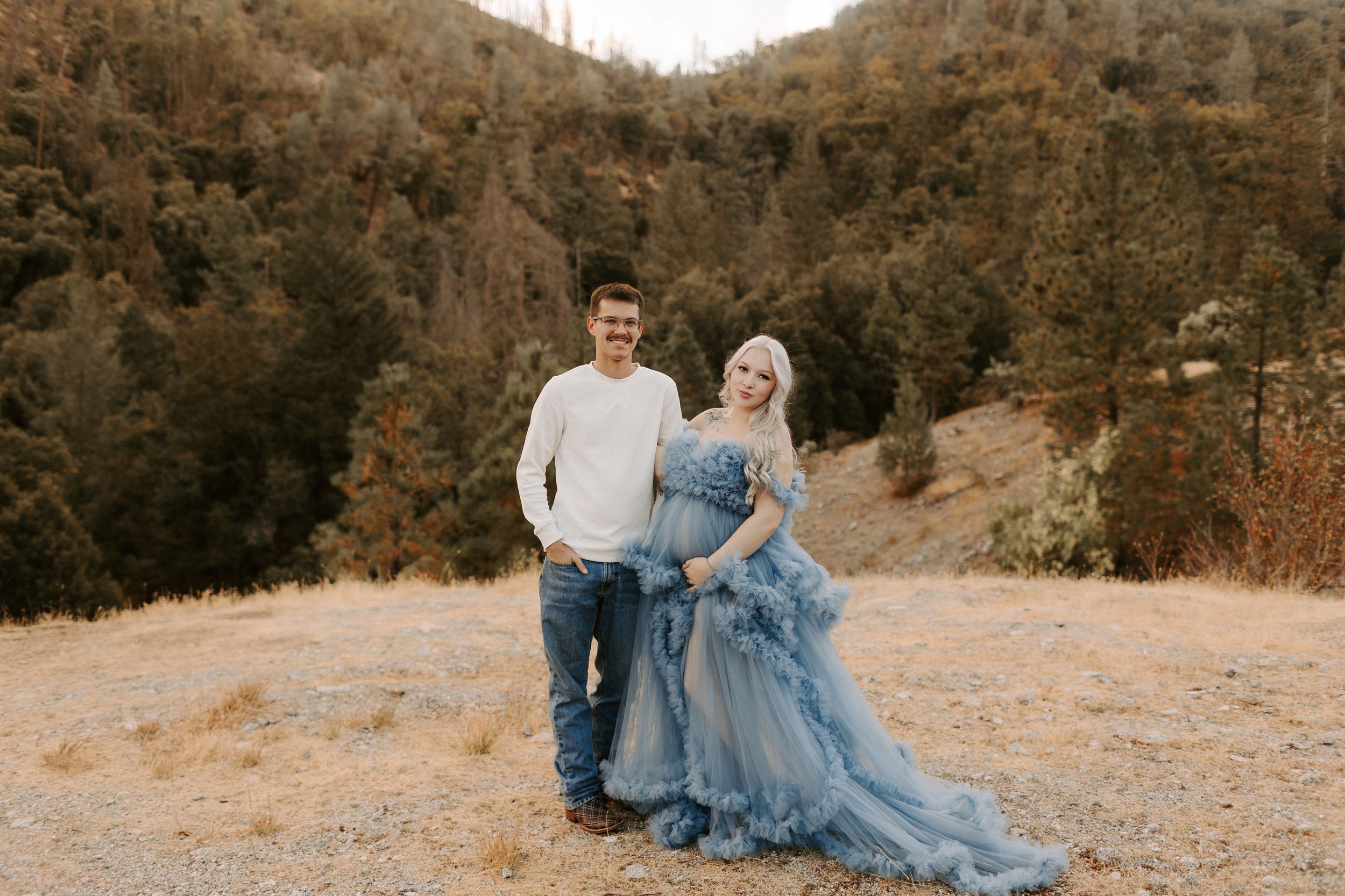 Romantic couple poses in field with mountains and golden light in background.