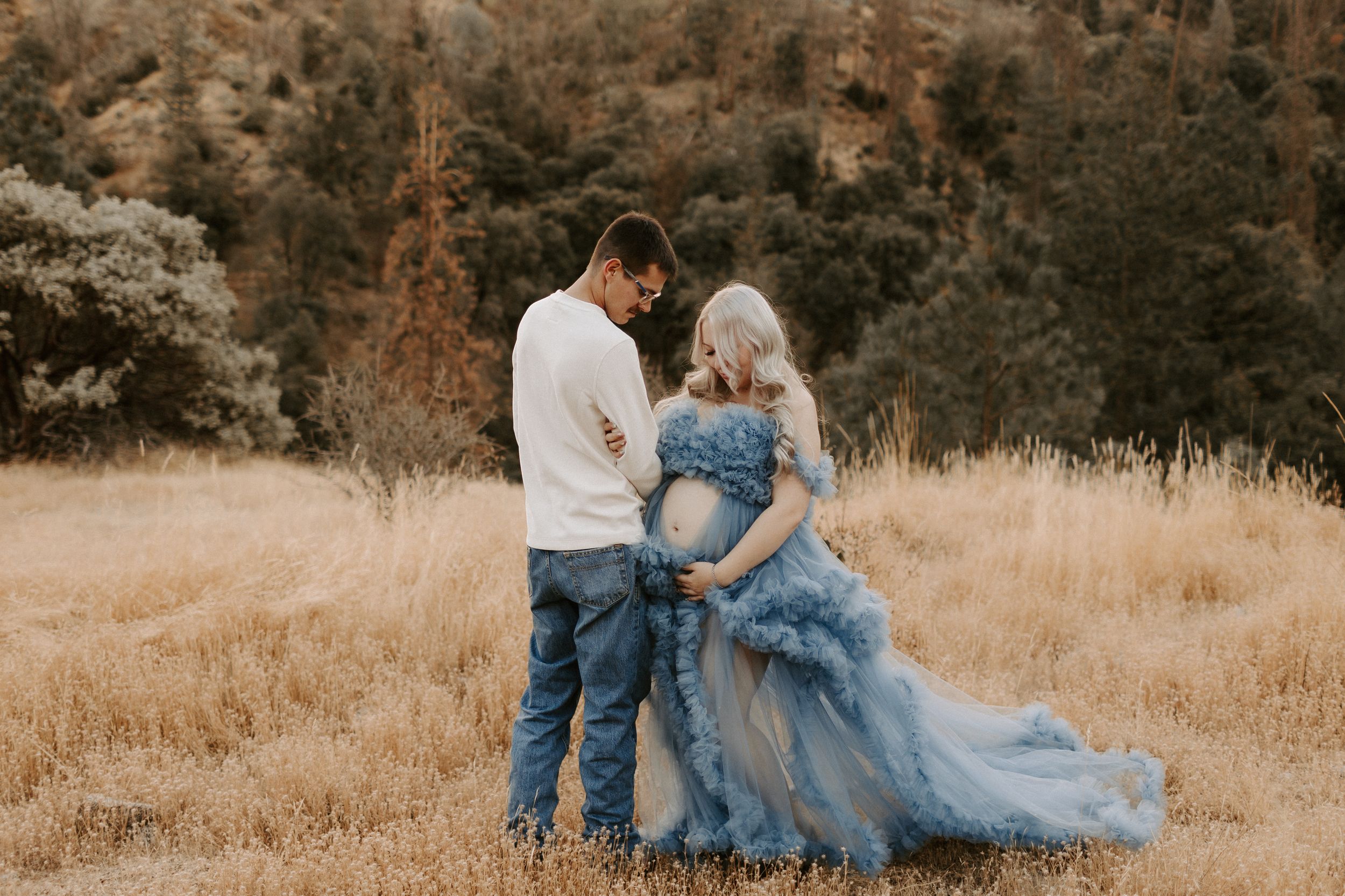 Couple embraces while wearing blue tulle gown and casual attire in autumn field.