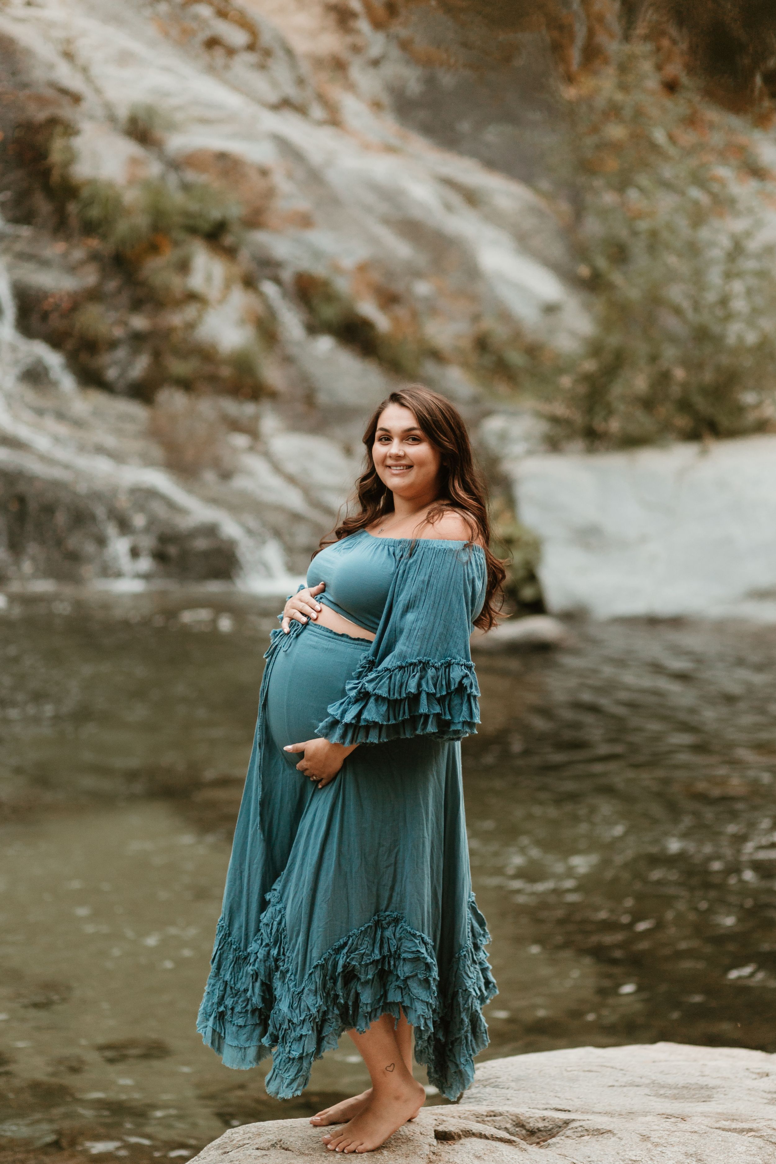 Maternity session by water featuring teal fringe dress and dramatic rock formations.