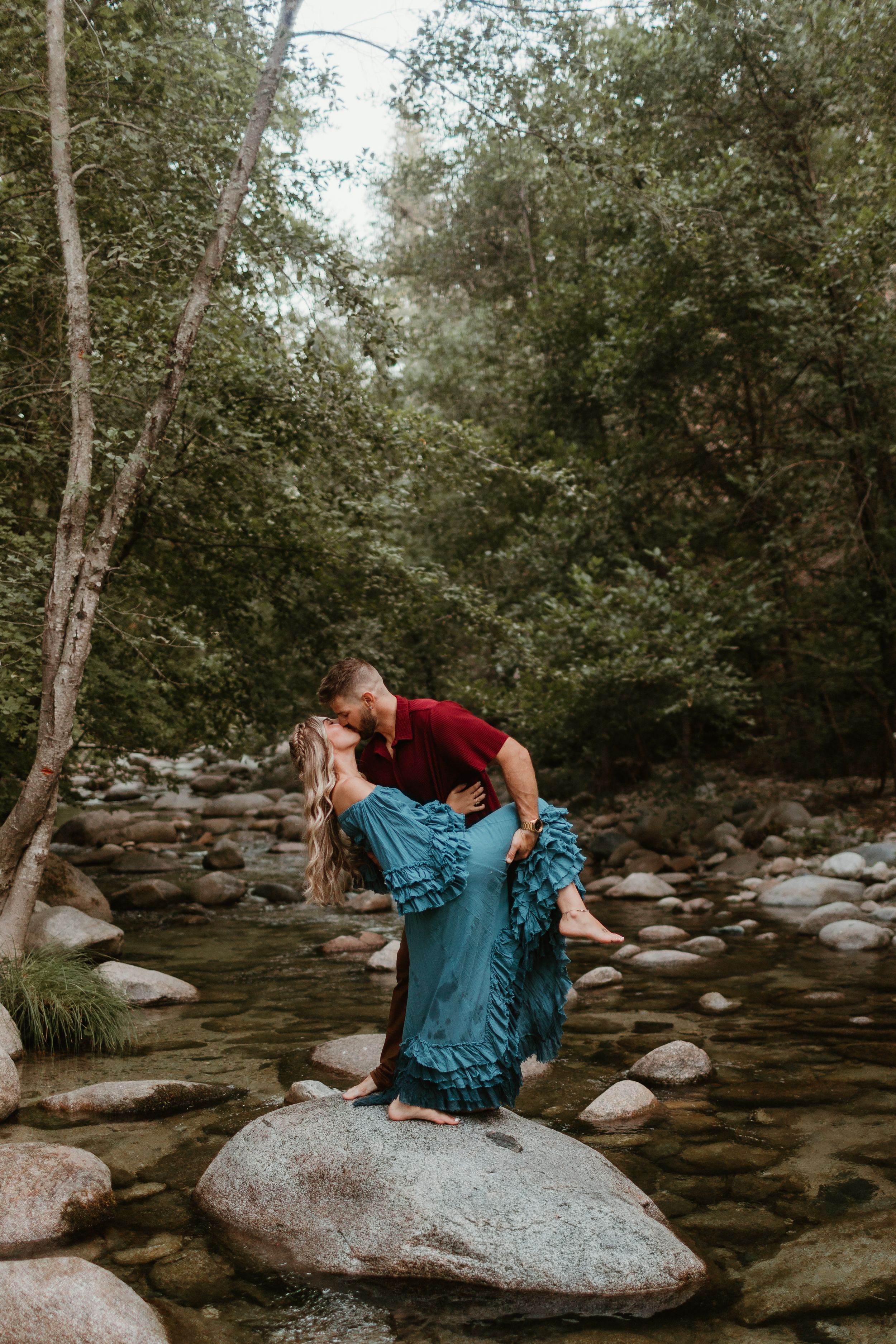 Couple in coordinated outfits sharing playful moments on large rocks by flowing stream.