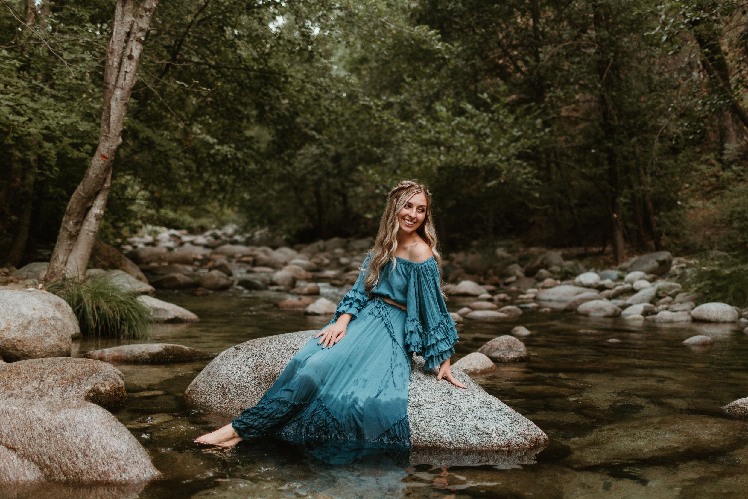 A teal dress flows elegantly against a rocky creek backdrop in a natural forest setting.