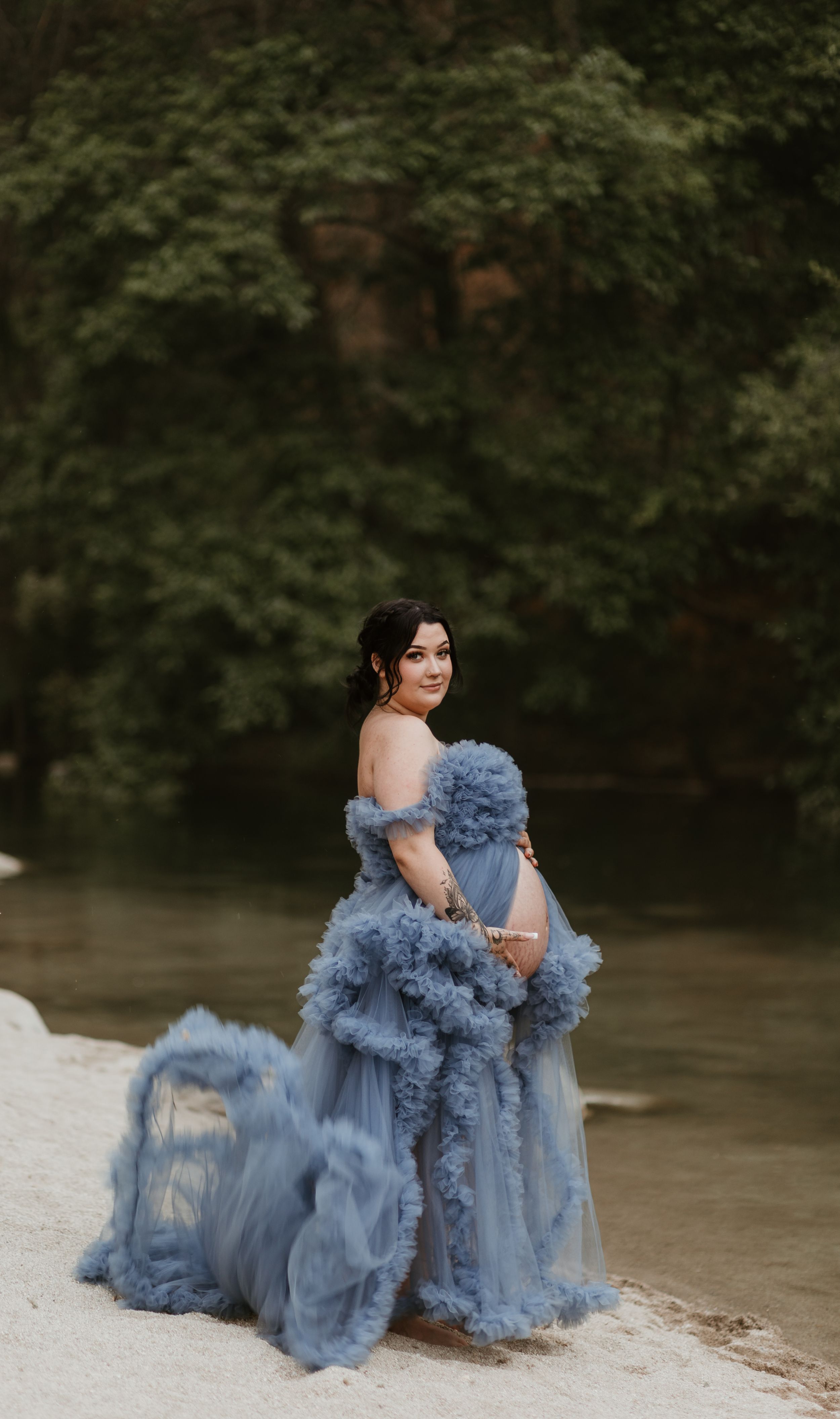 Dramatic blue tulle dress flows across rocky riverbank during outdoor photo session.