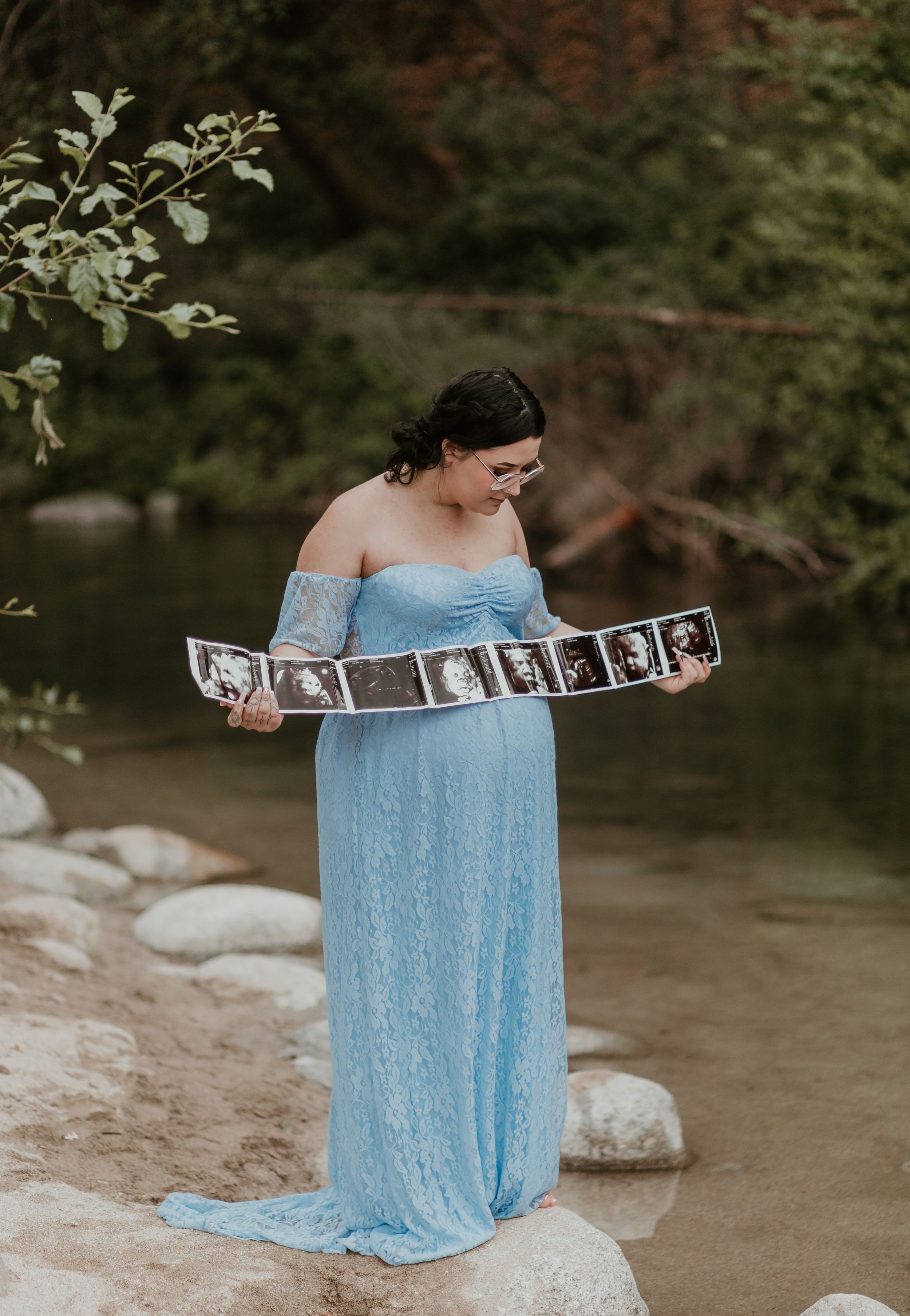 Person in a light blue off-shoulder dress holding photographs while sitting by a stream.