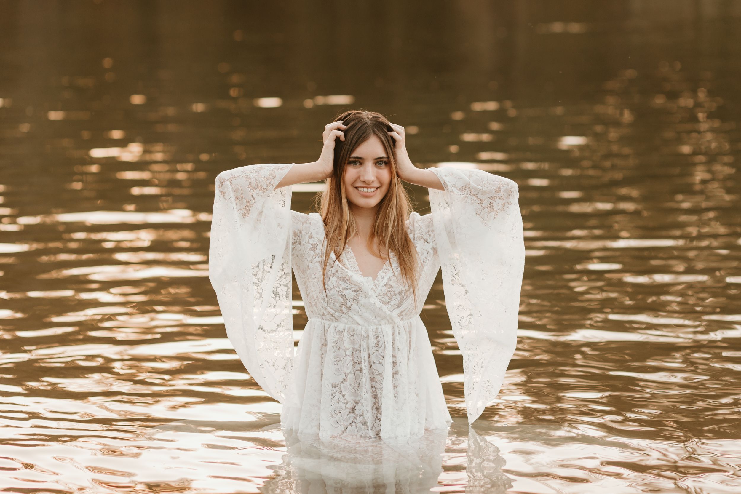 Flowing white lace dress creates dreamy silhouette against sunlit water backdrop.