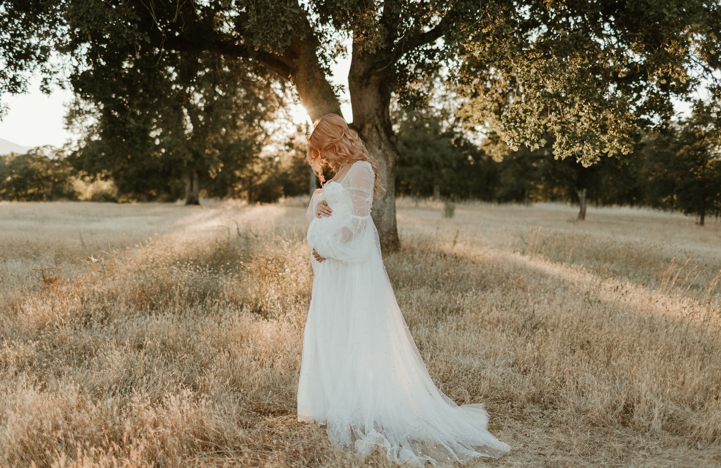 A flowing white gown trails beautifully across dried grass in a sun-lit meadow.