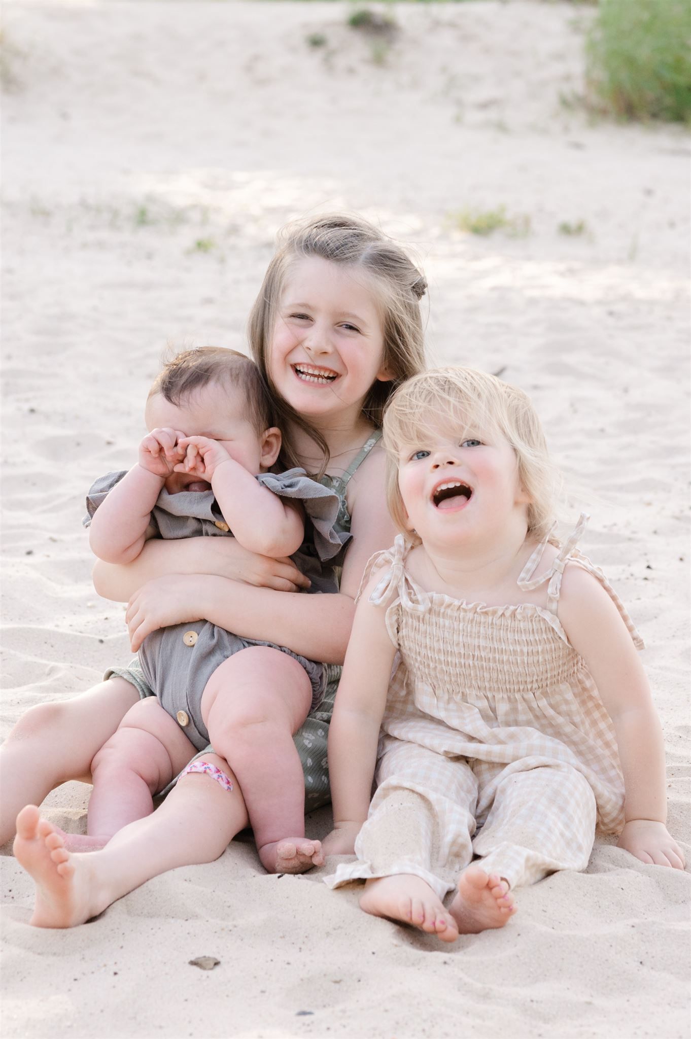 Three children sitting on sand laughing and playing together in casual summer outfits.